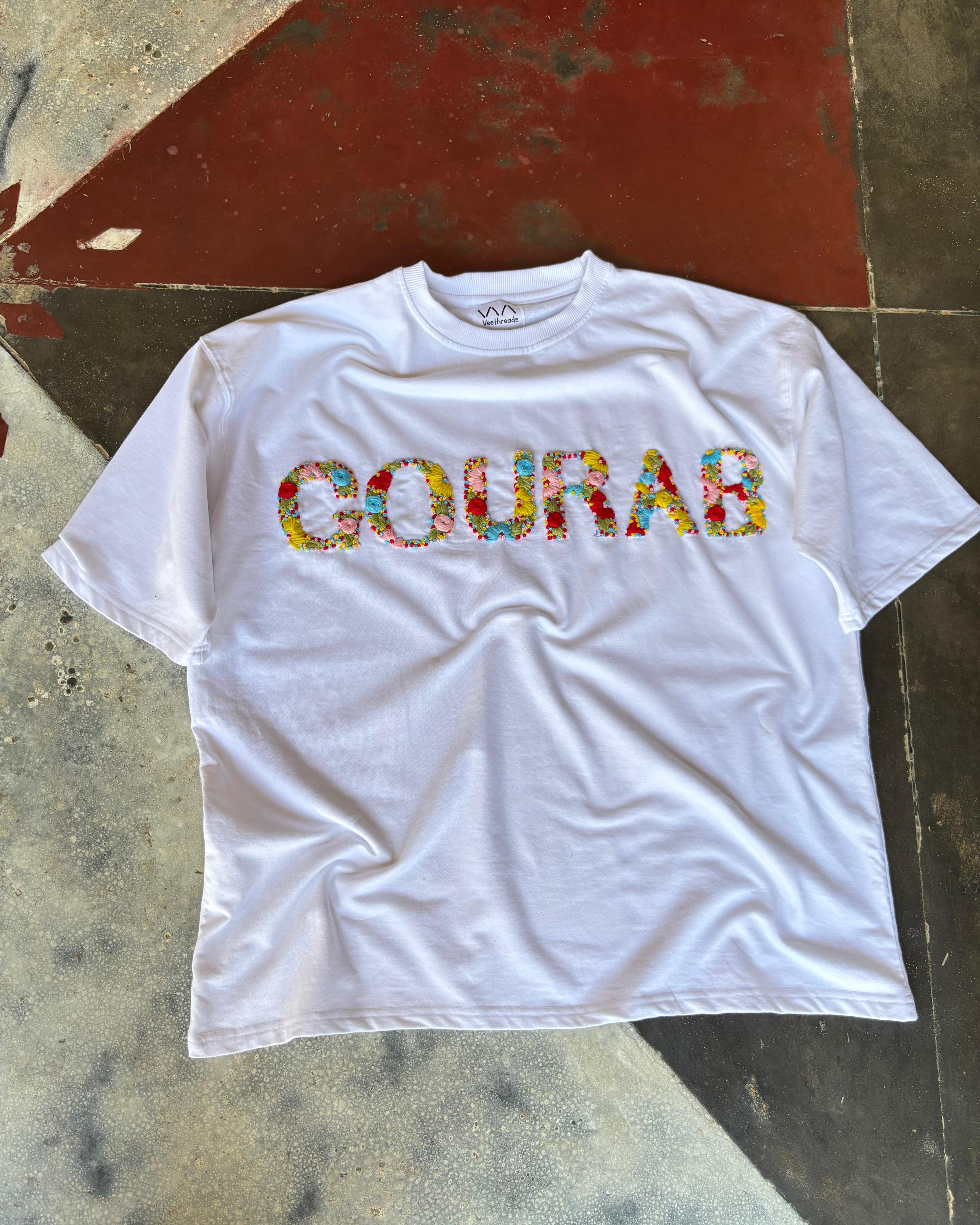 Oversized embroidered shirt laid flat on a concrete surface, showing detailed name stitching near the heart.