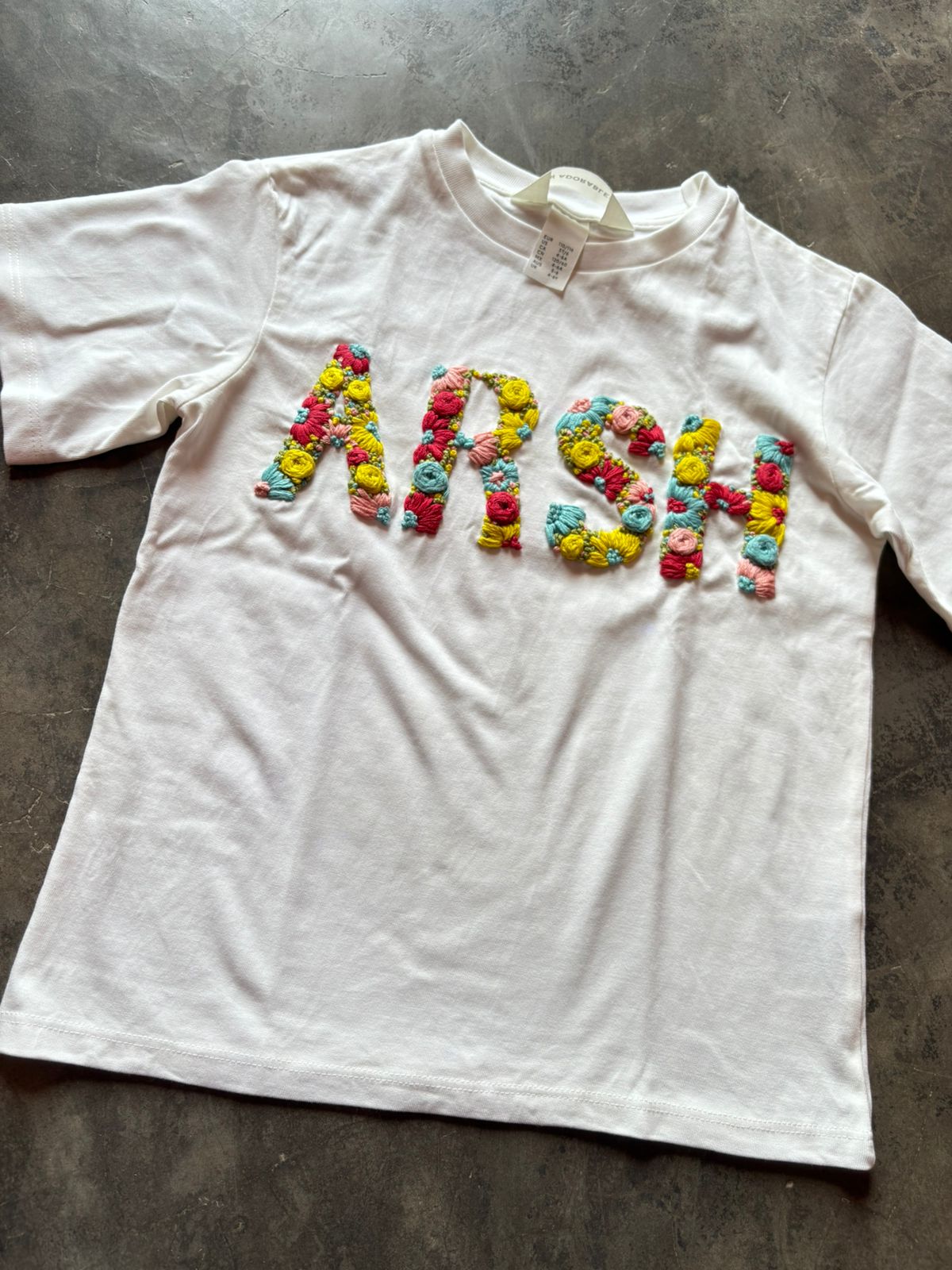 White t-shirt with customised embroidery spelling 'ARSH' on a gray surface