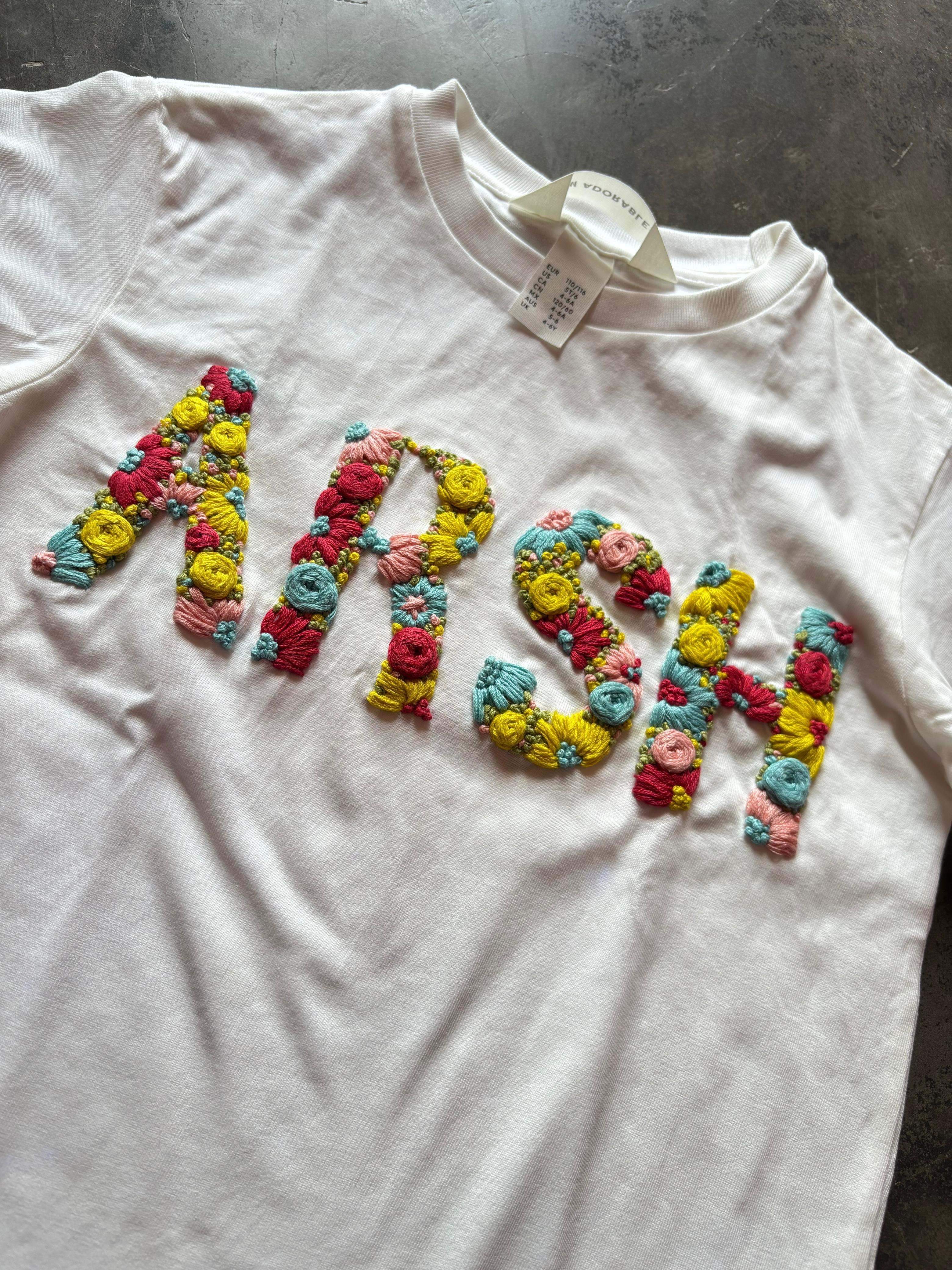 White t-shirt with colorful floral embroidered letters spelling 'MRS.' on a gray surface