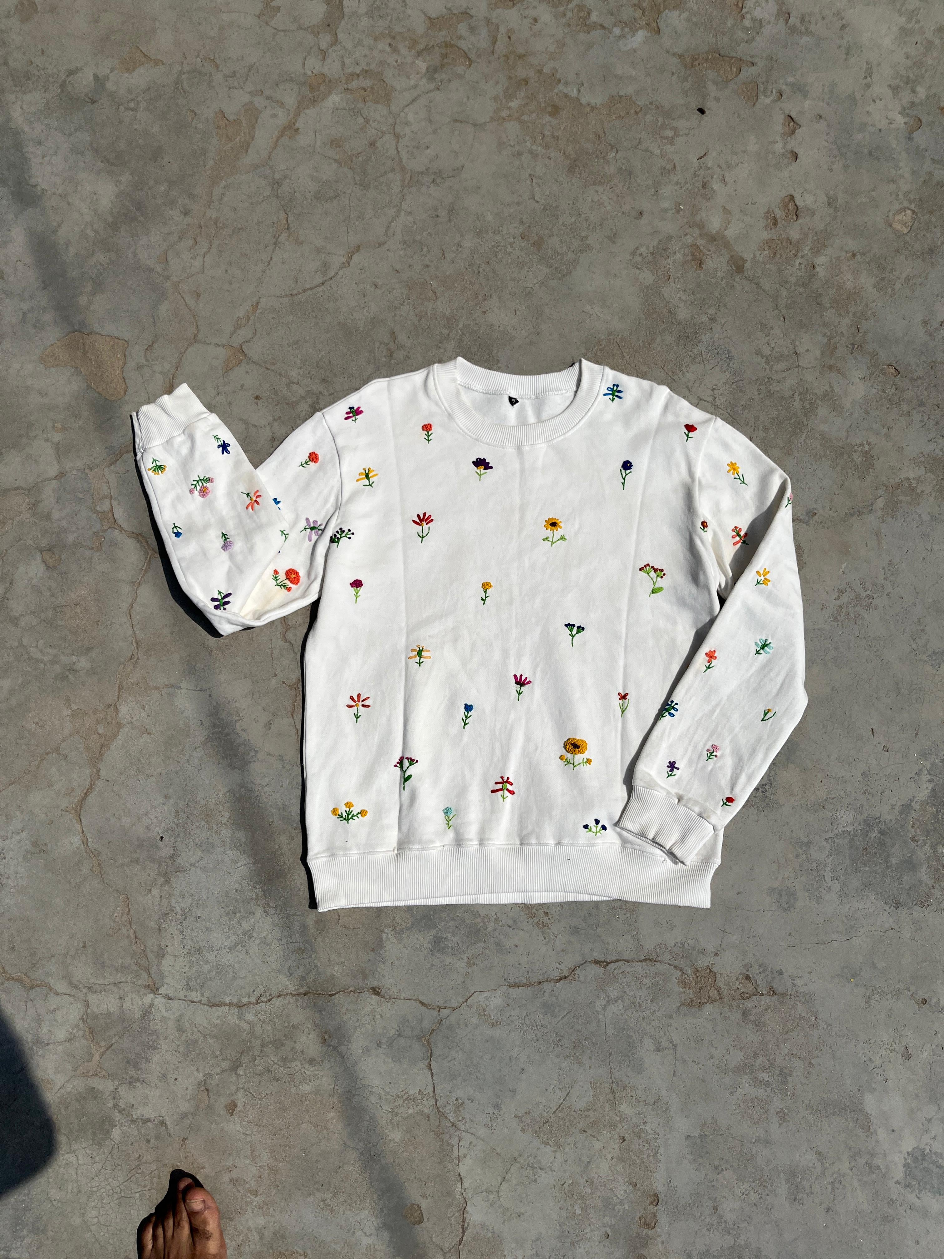 White flower embroidered sweatshirt with colorful patterns on a gray concrete floor