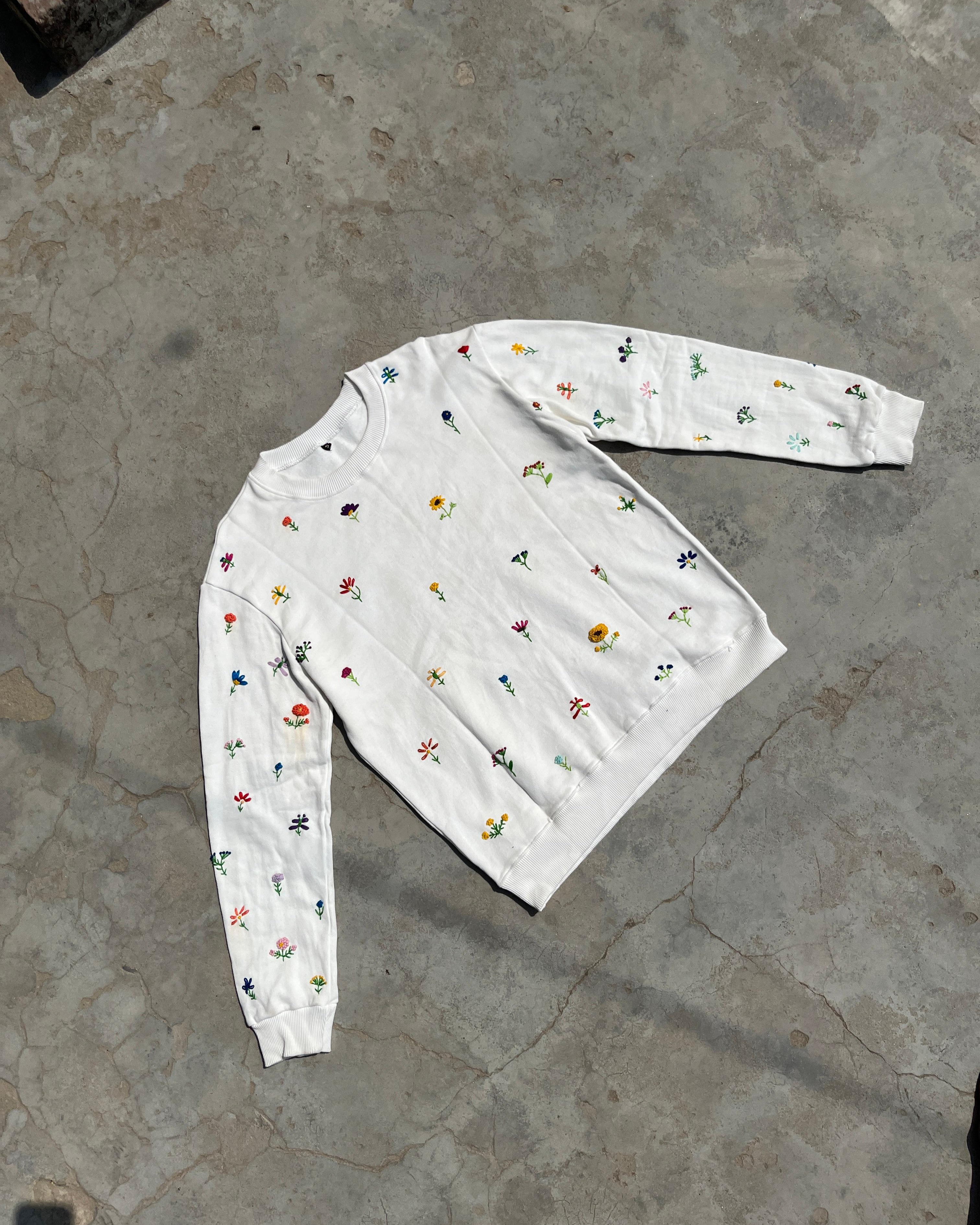 White long-sleeve sweatshirt with colorful floral hand embroidered patterns on a concrete floor