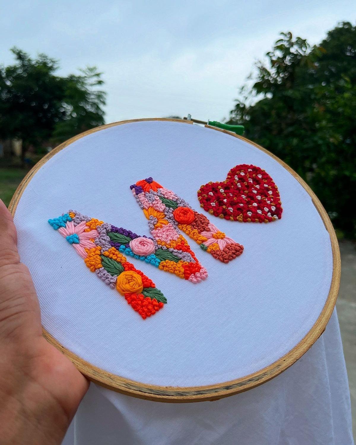 Embroidery hoop with colorful floral design and heart shape held outdoors.