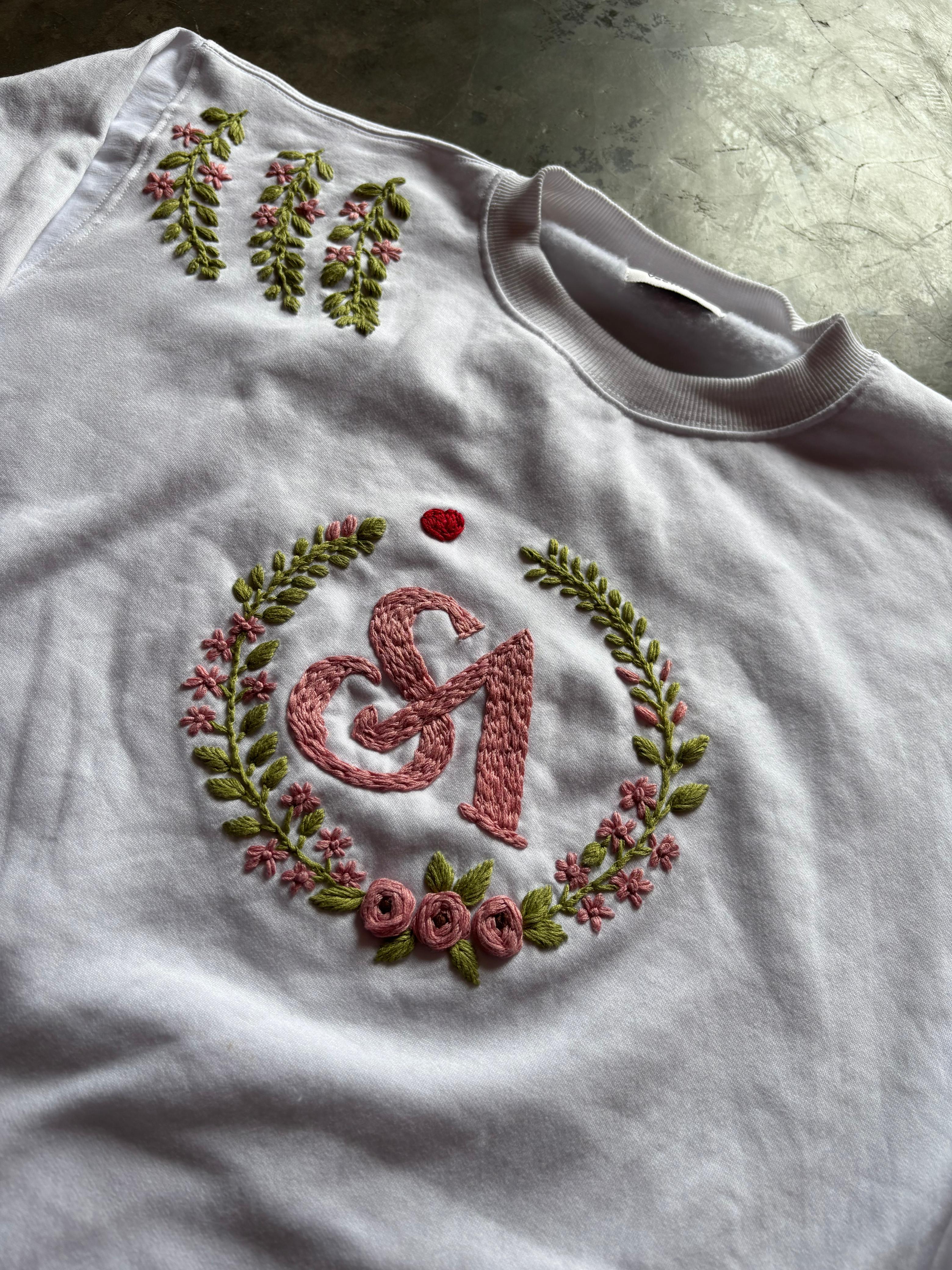 White shirt with embroidered design of a letter 'S' and floral elements on a textured surface.