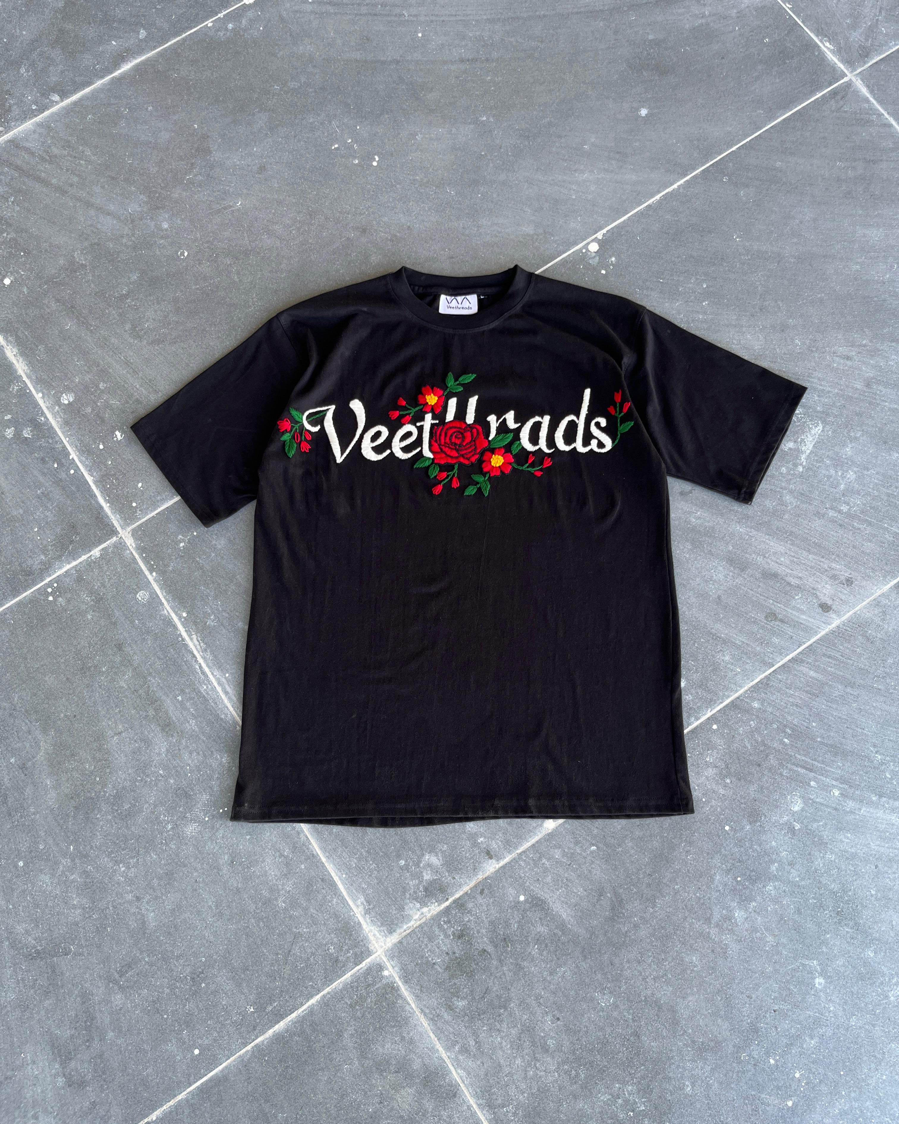 Black t-shirt with 'Veet Vrads' text and floral design on a gray tiled floor.