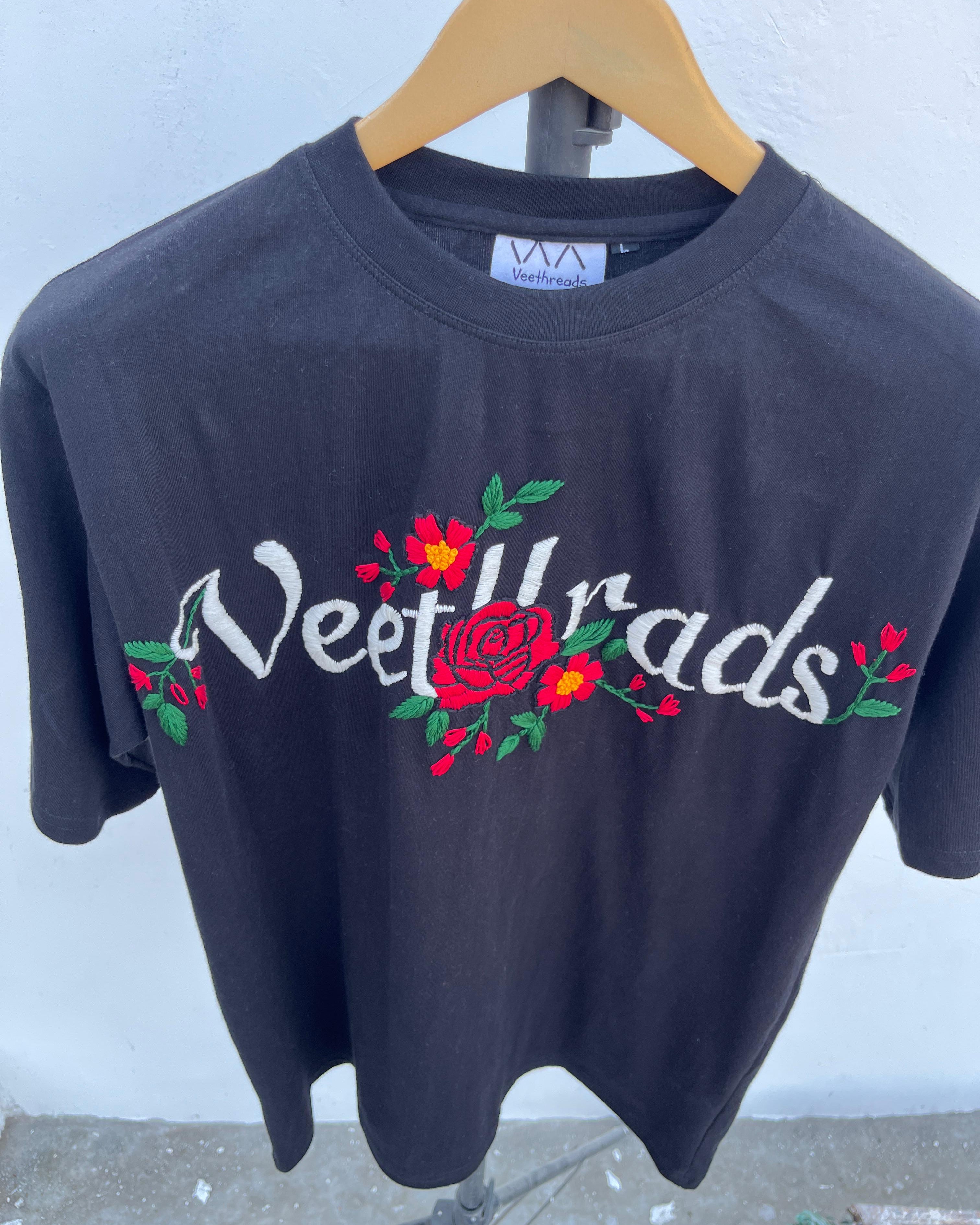 Black t-shirt with 'Veet Wrads' text and floral designs on a hanger against a light blue wall.