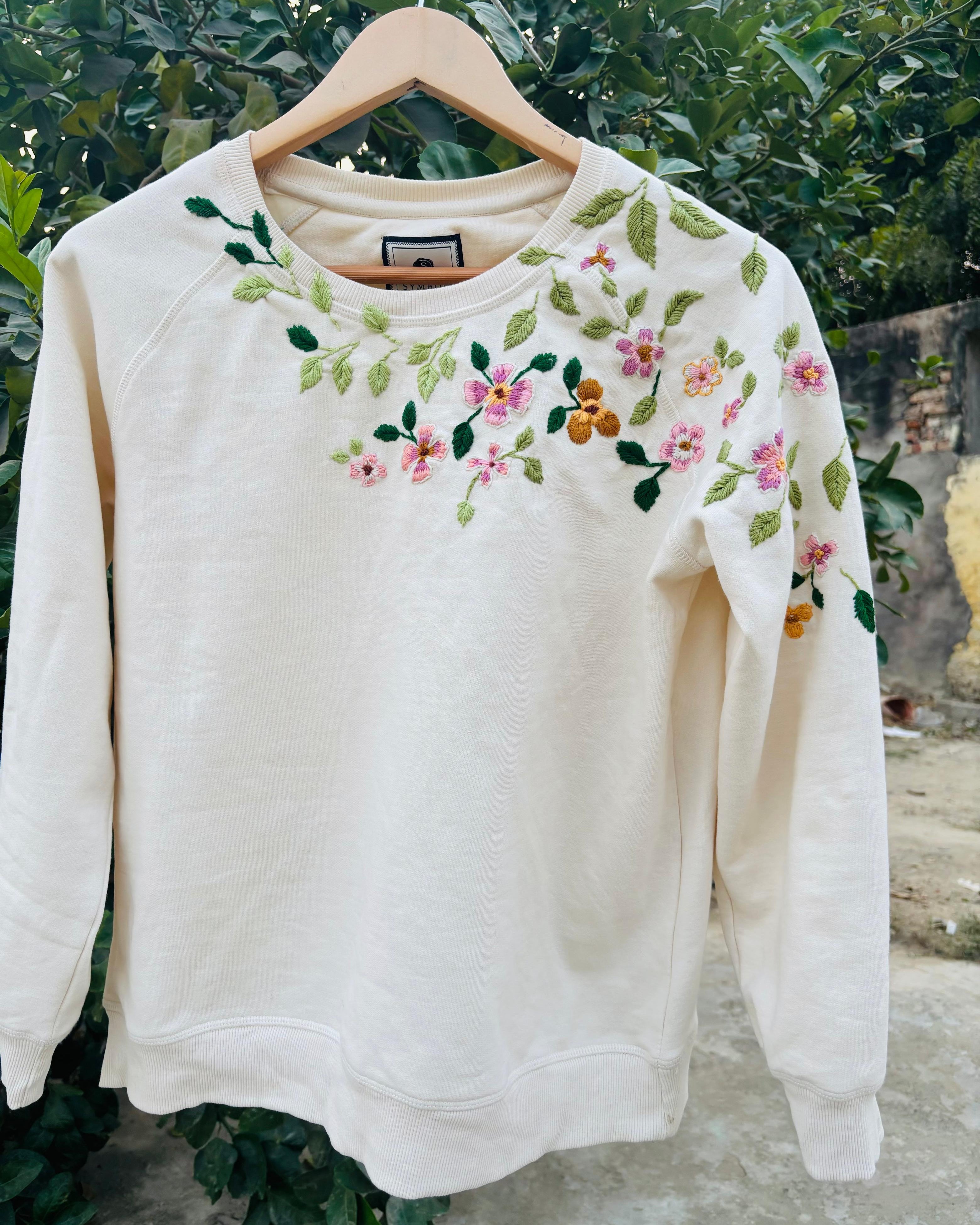 White sweatshirt with floral embroidery on a wooden hanger against a natural background