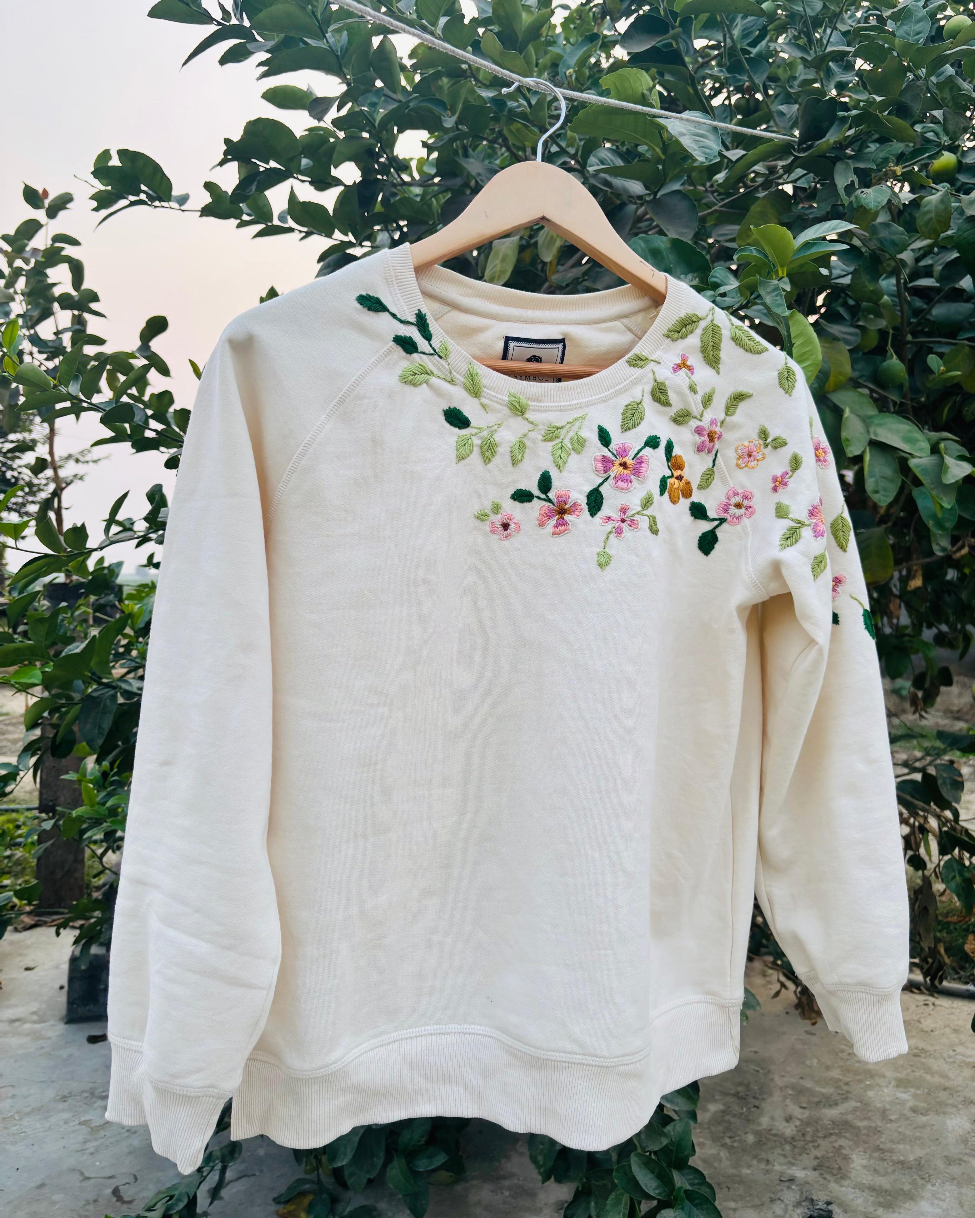 White sweatshirt with floral embroidery on a hanger against a green foliage background