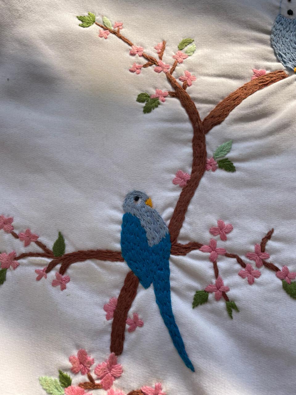 Embroidered scene with a blue parrot on a branch with pink flowers on a white fabric background.