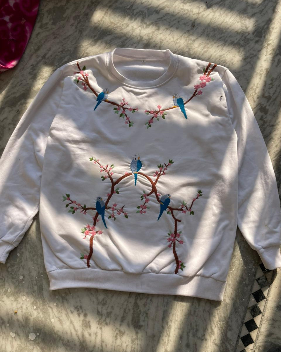 White sweater with embroidered birds and branches on a textured surface
