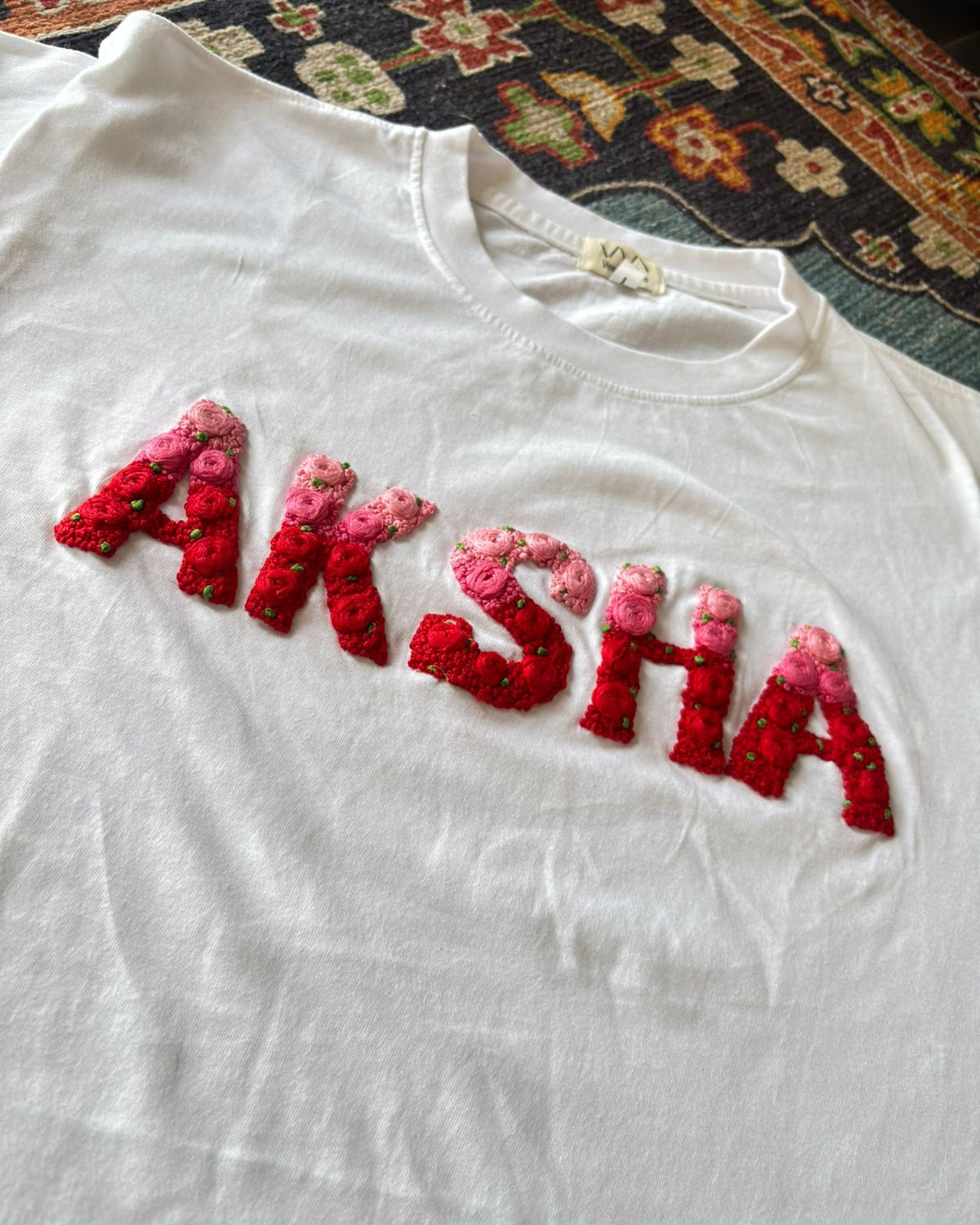 White t-shirt with 'AKSHA' embroidered in red and pink on a patterned rug.