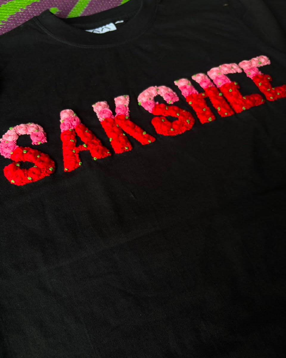 Black sweatshirt with 'SAKSHIM' text made of red and pink sequins on a colorful fabric background.