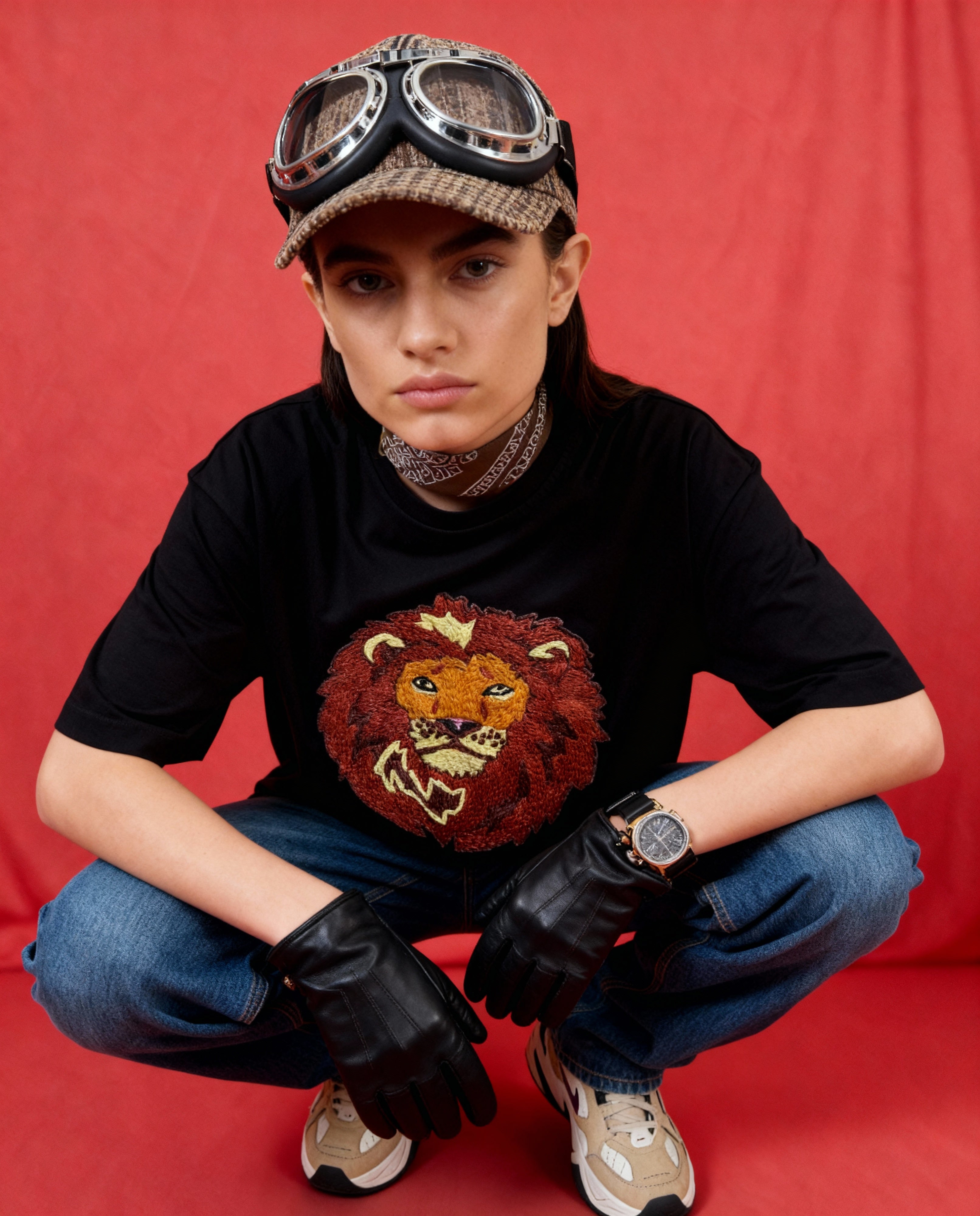 Person wearing a black t-shirt with a lion graphic, jeans, gloves, and a cap with goggles on a red background
