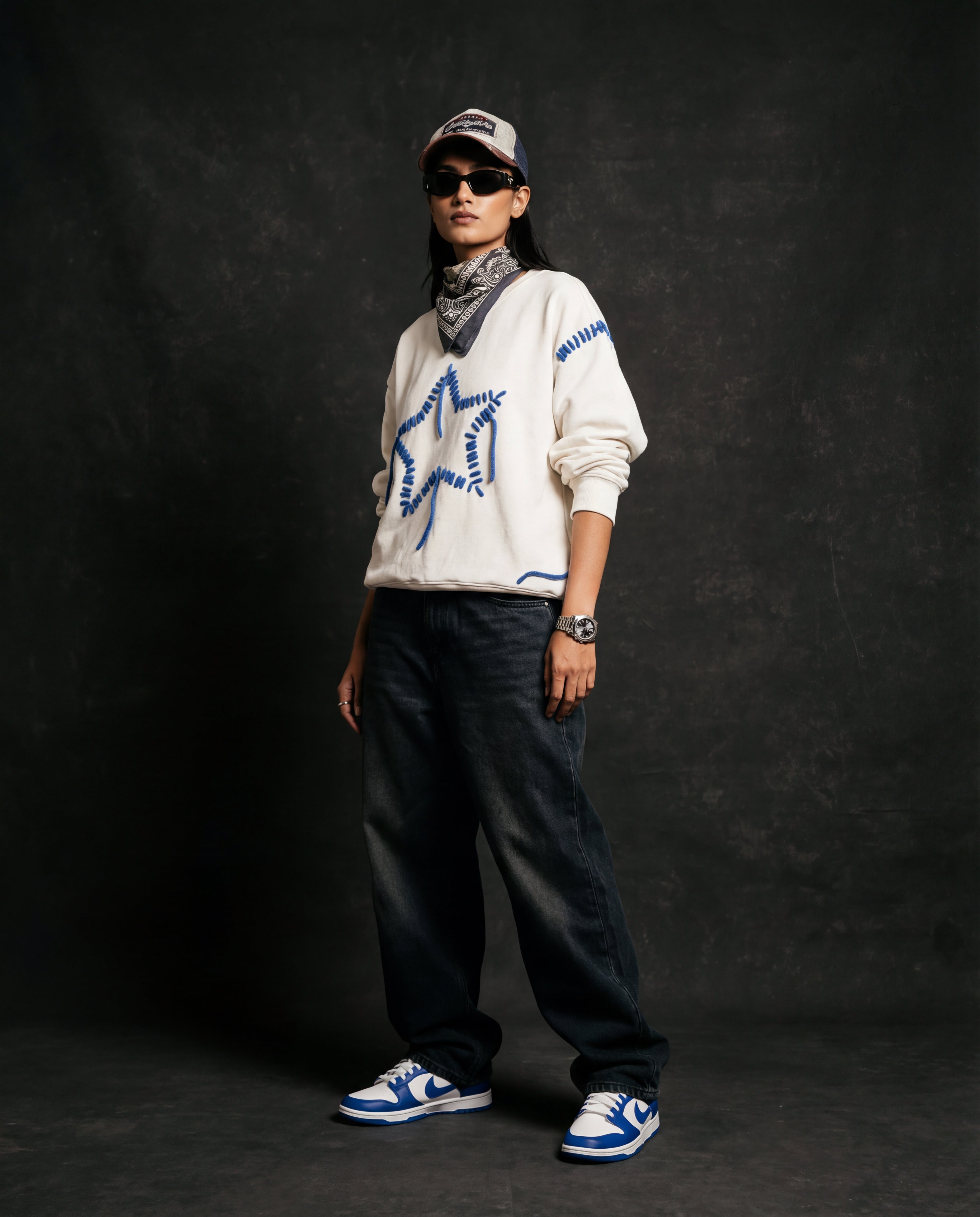 Person wearing a white sweatshirt with embroidered blue star design, black pants, and blue sneakers against a dark background