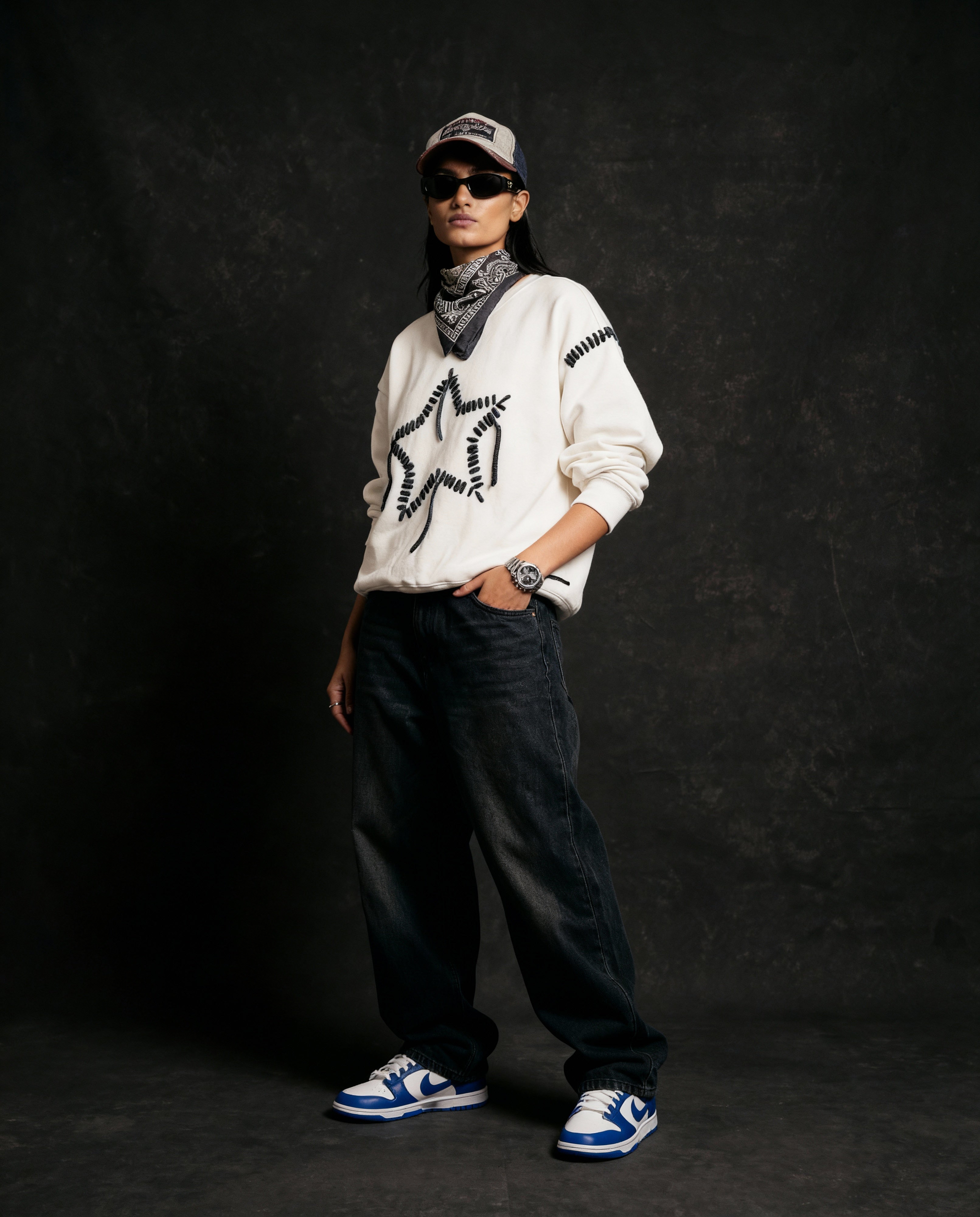 Person wearing a white sweater with black patterns, dark pants, and blue sneakers against a black background