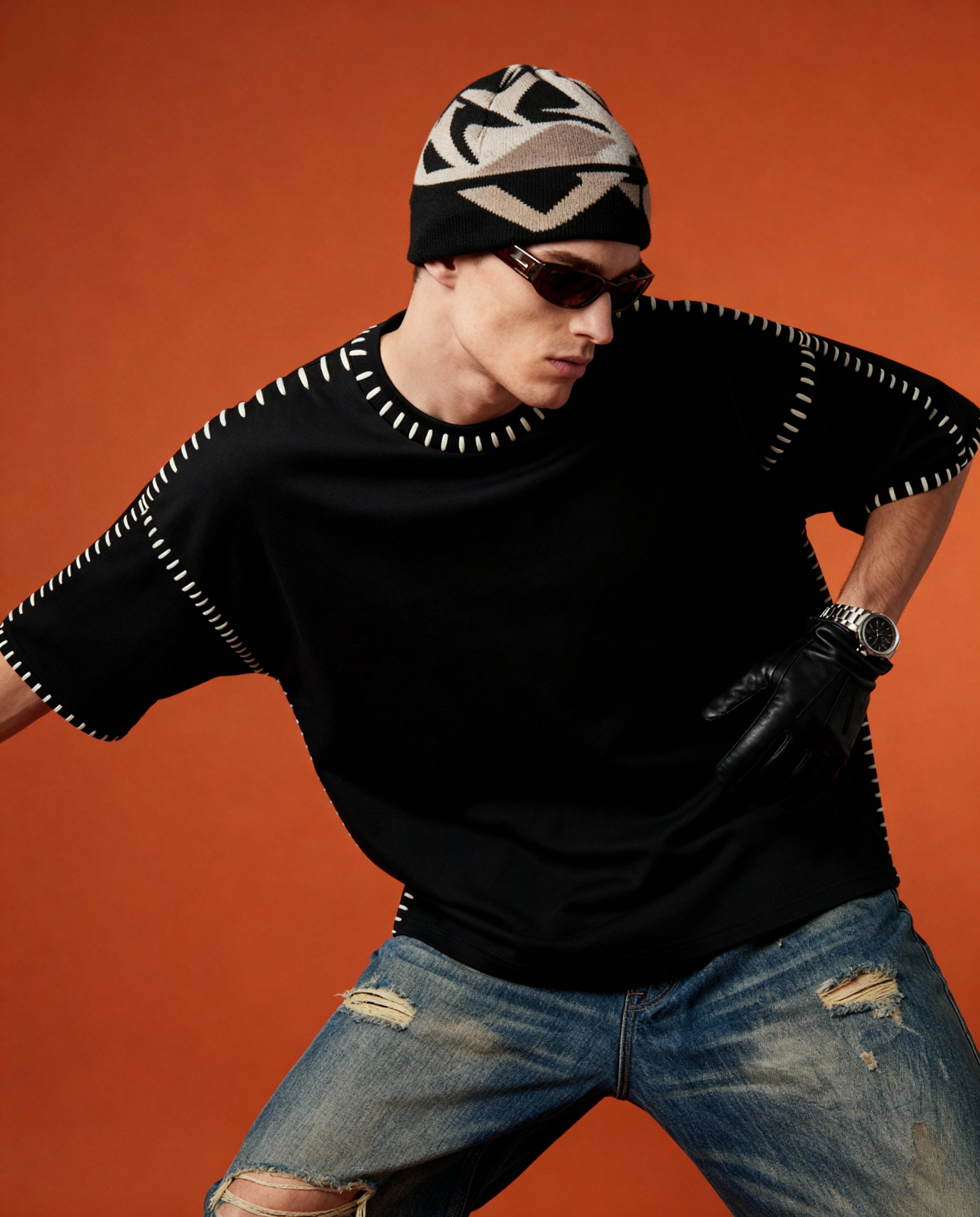 Person wearing a black outfit with white accents, sunglasses, and a patterned beanie against an orange background