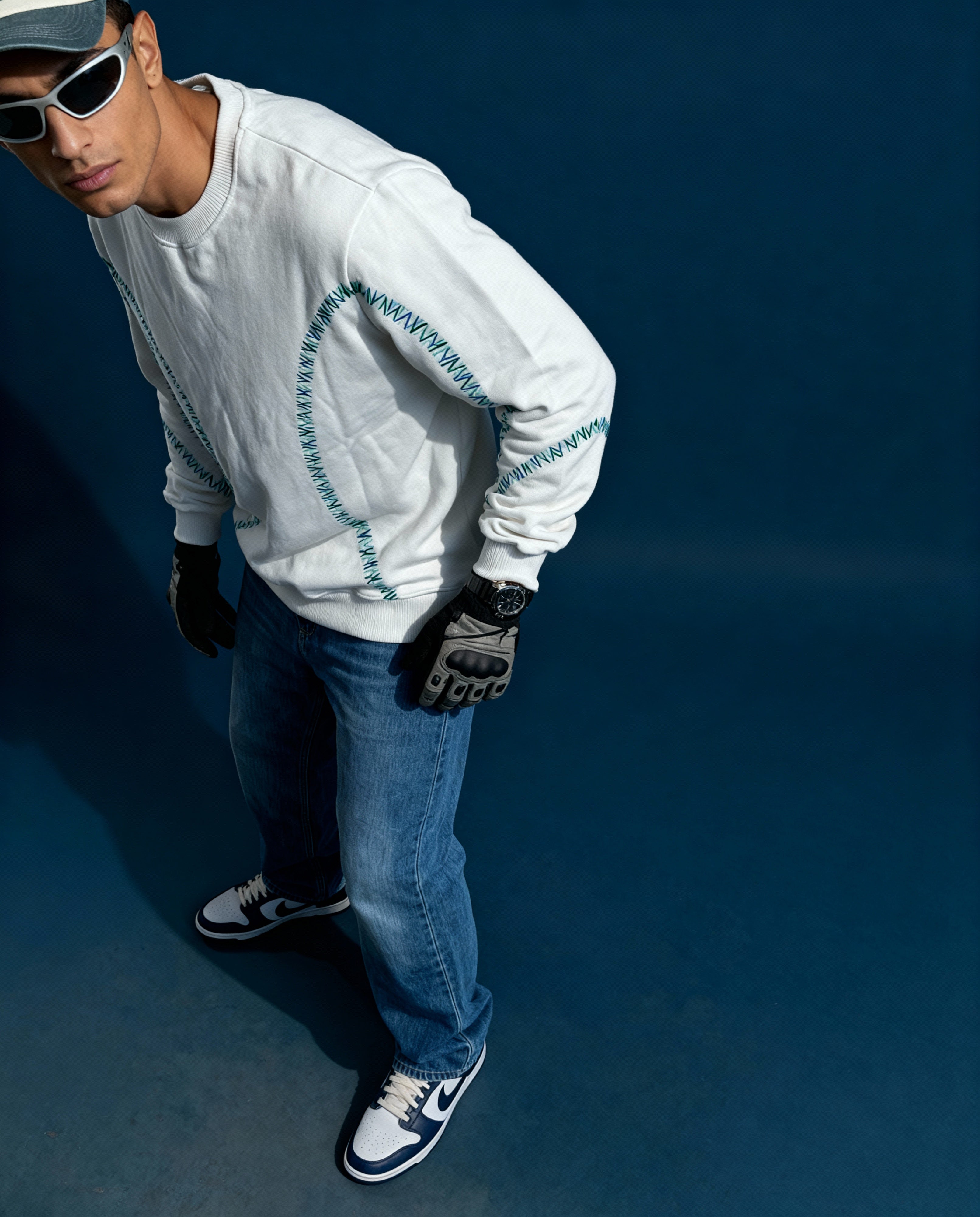 Person wearing a white sweater with blue accents, blue jeans, and black shoes on a dark background