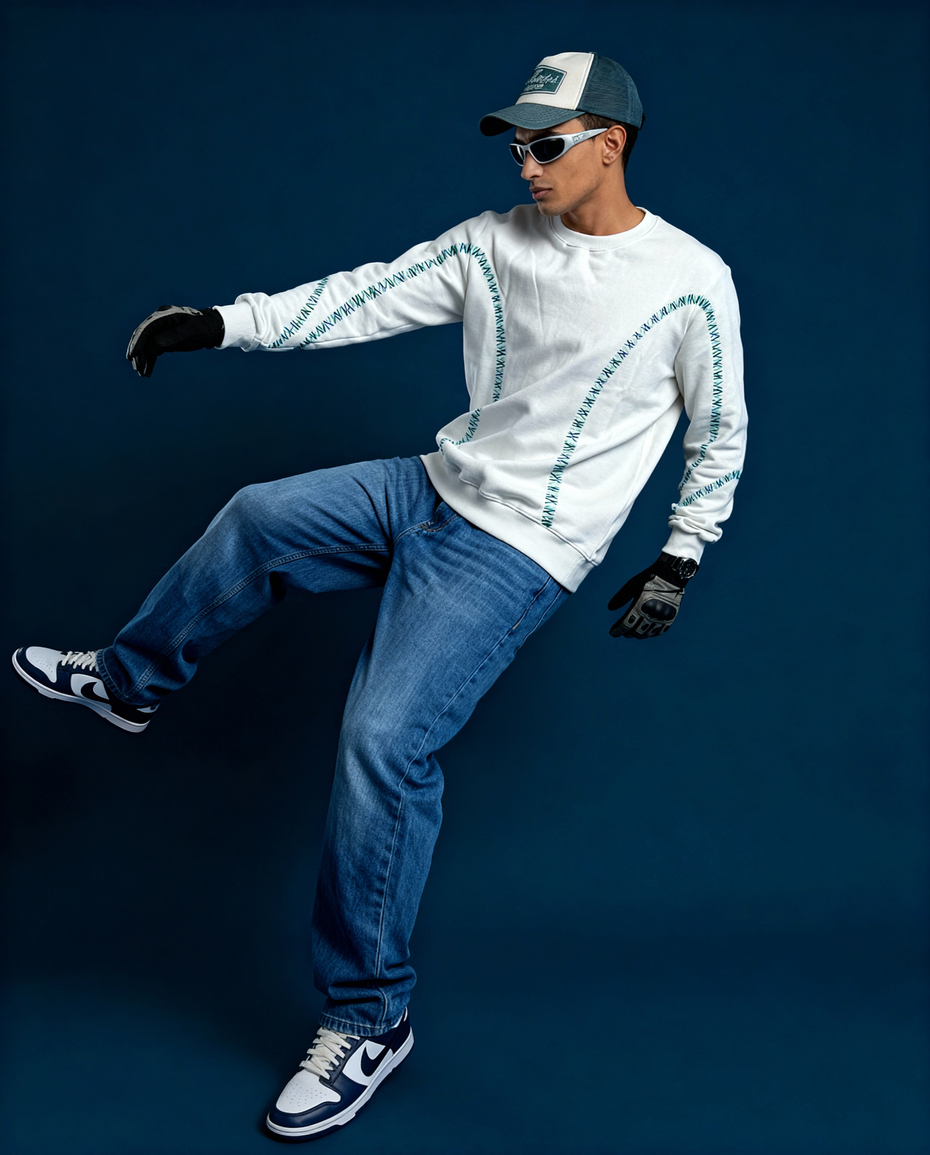 Person wearing a white sweatshirt with blue accents, blue jeans, black gloves, and a cap on a dark background