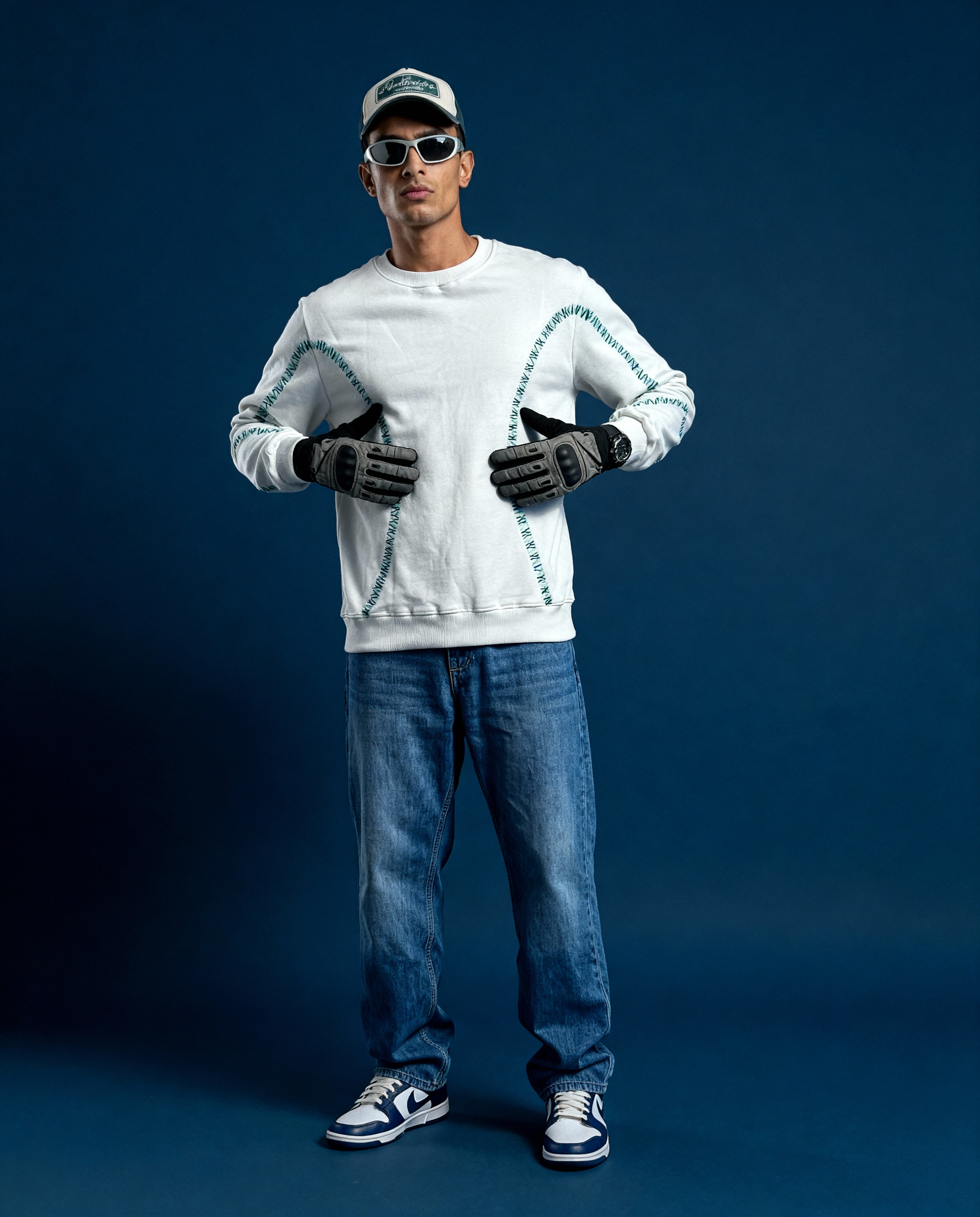 Man wearing a white sweater with a unique design, blue jeans, and black shoes against a dark blue background.