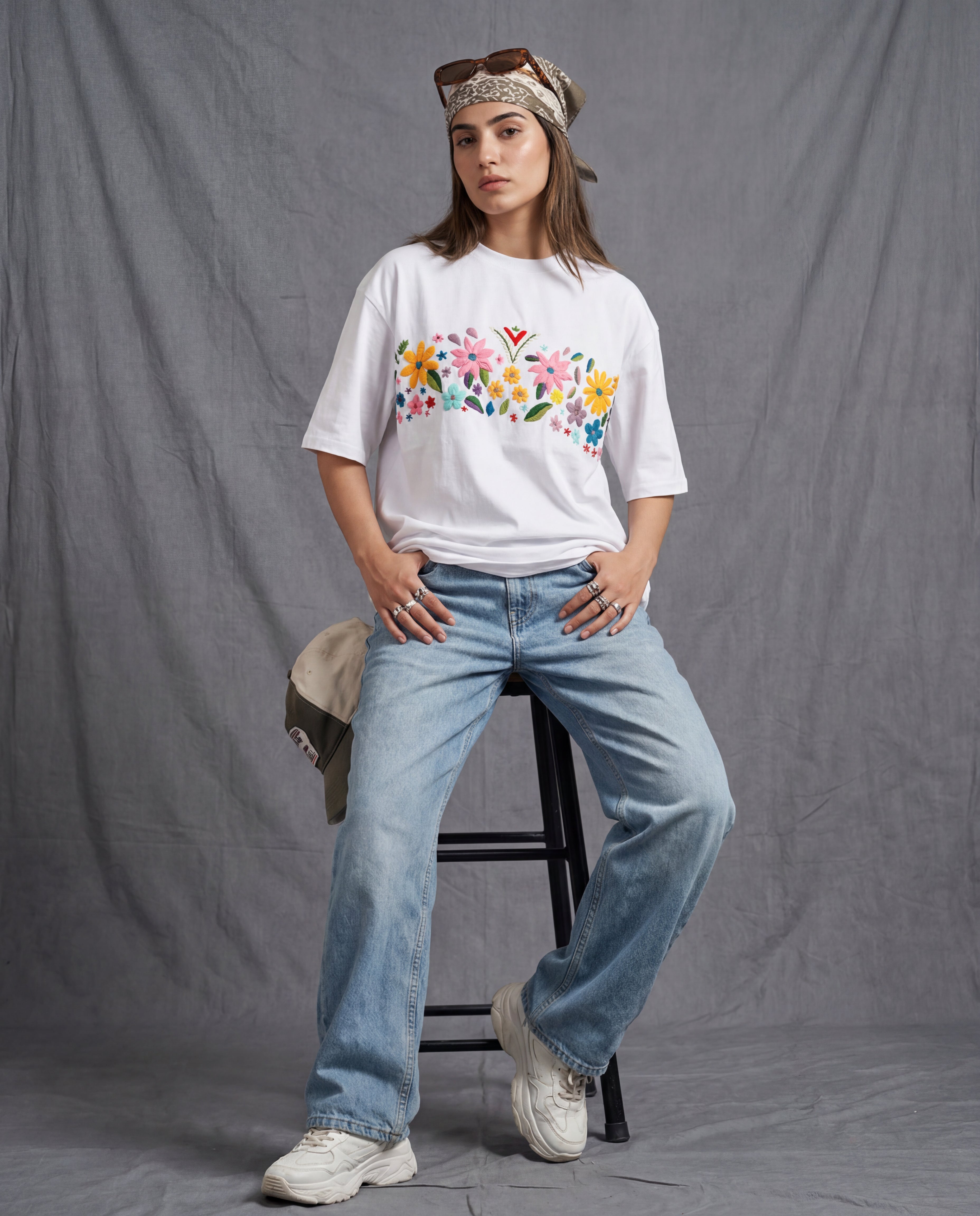 Person wearing a white t-shirt with colorful floral hand embroidered design and blue jeans, standing against a gray background.