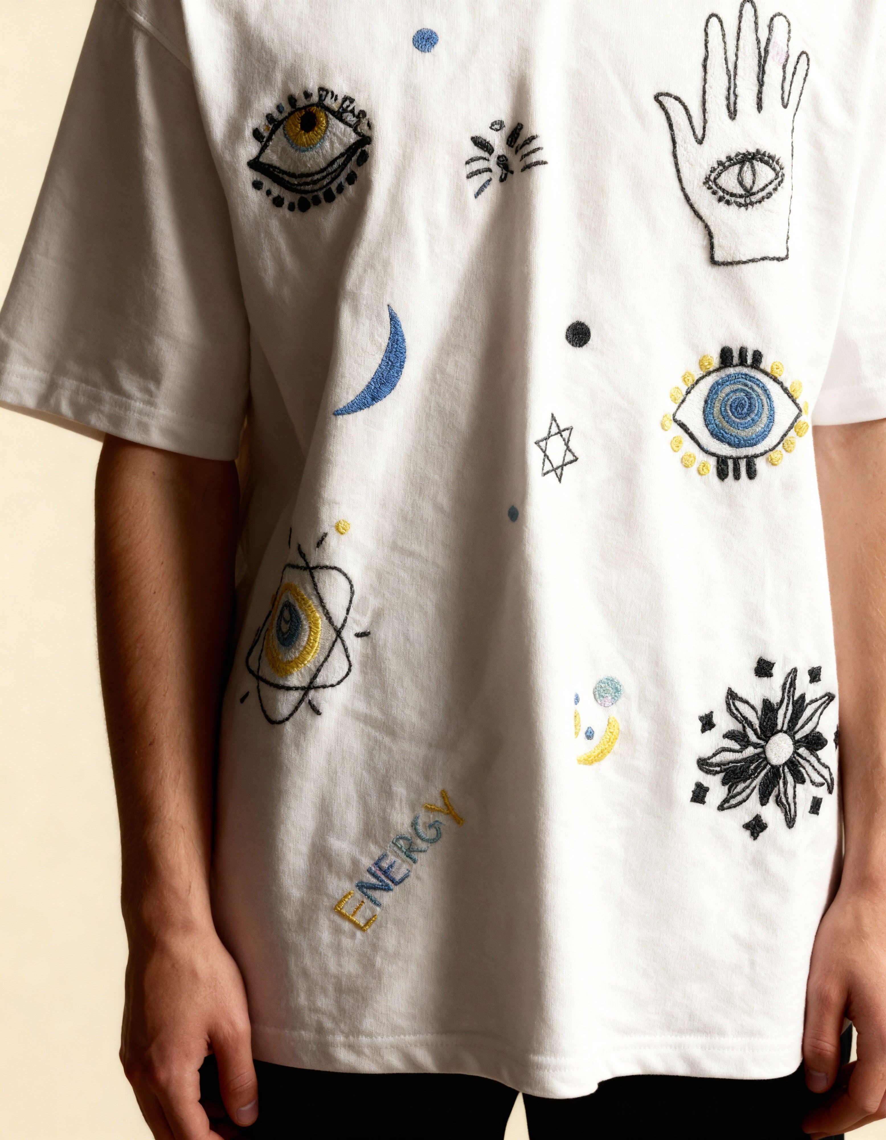 White t-shirt with eye and hand embroidered designs on a beige background