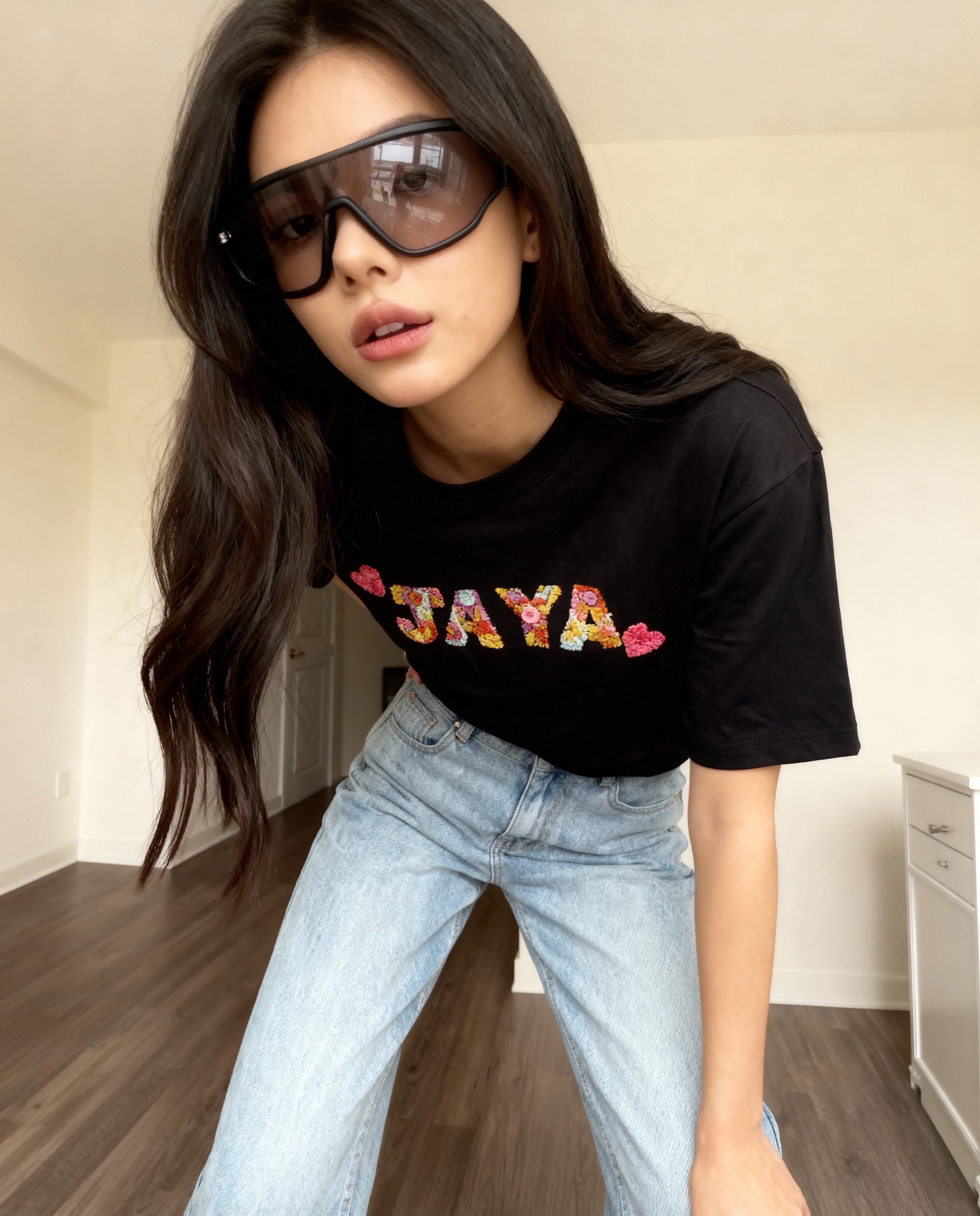 Person wearing a black t-shirt with 'JAYA' hand embroidered in colorful letters, sunglasses, and light blue jeans indoors.