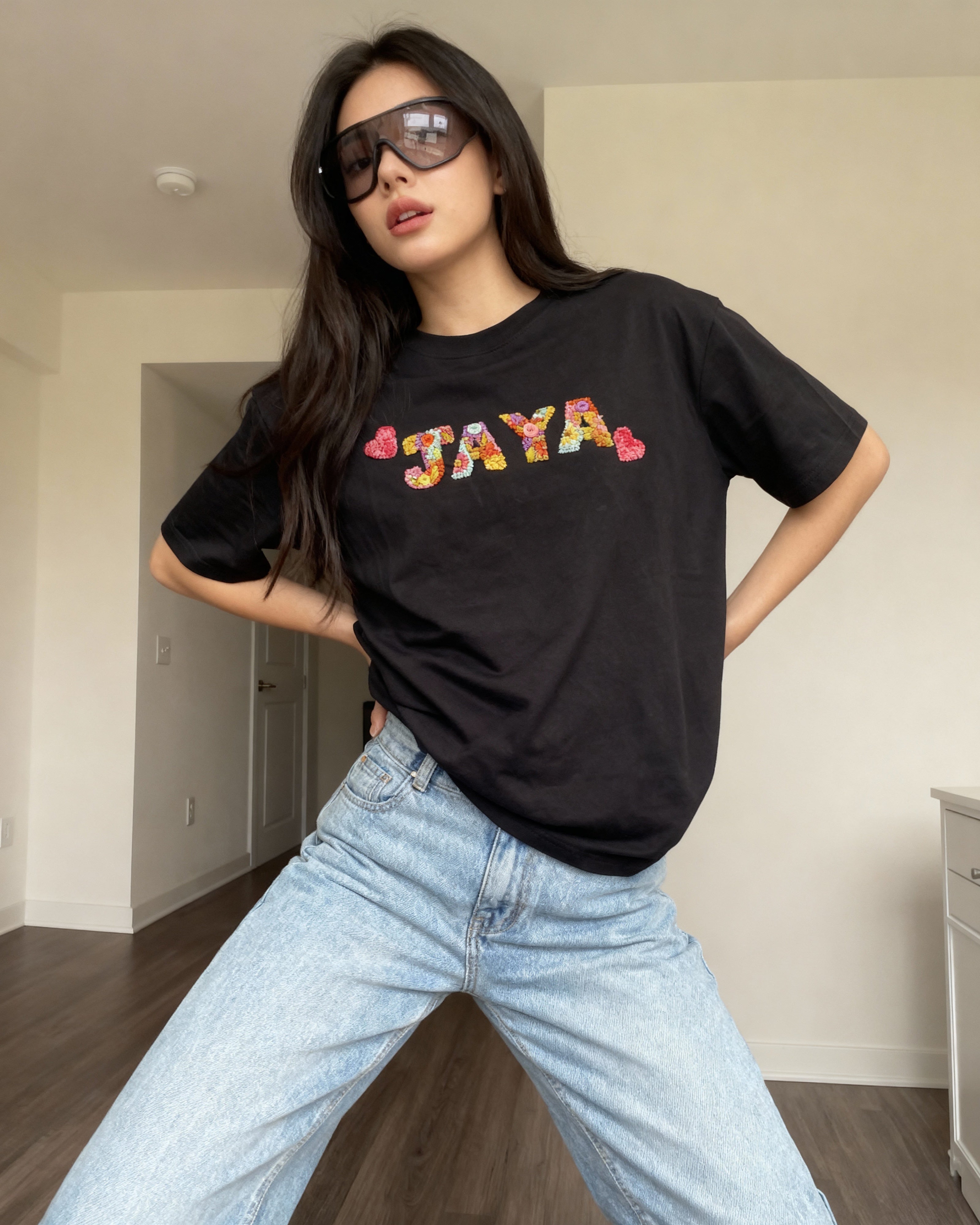 Person wearing a black t-shirt with 'JAYA' hand embroidered in colorful letters, standing in an indoor setting.