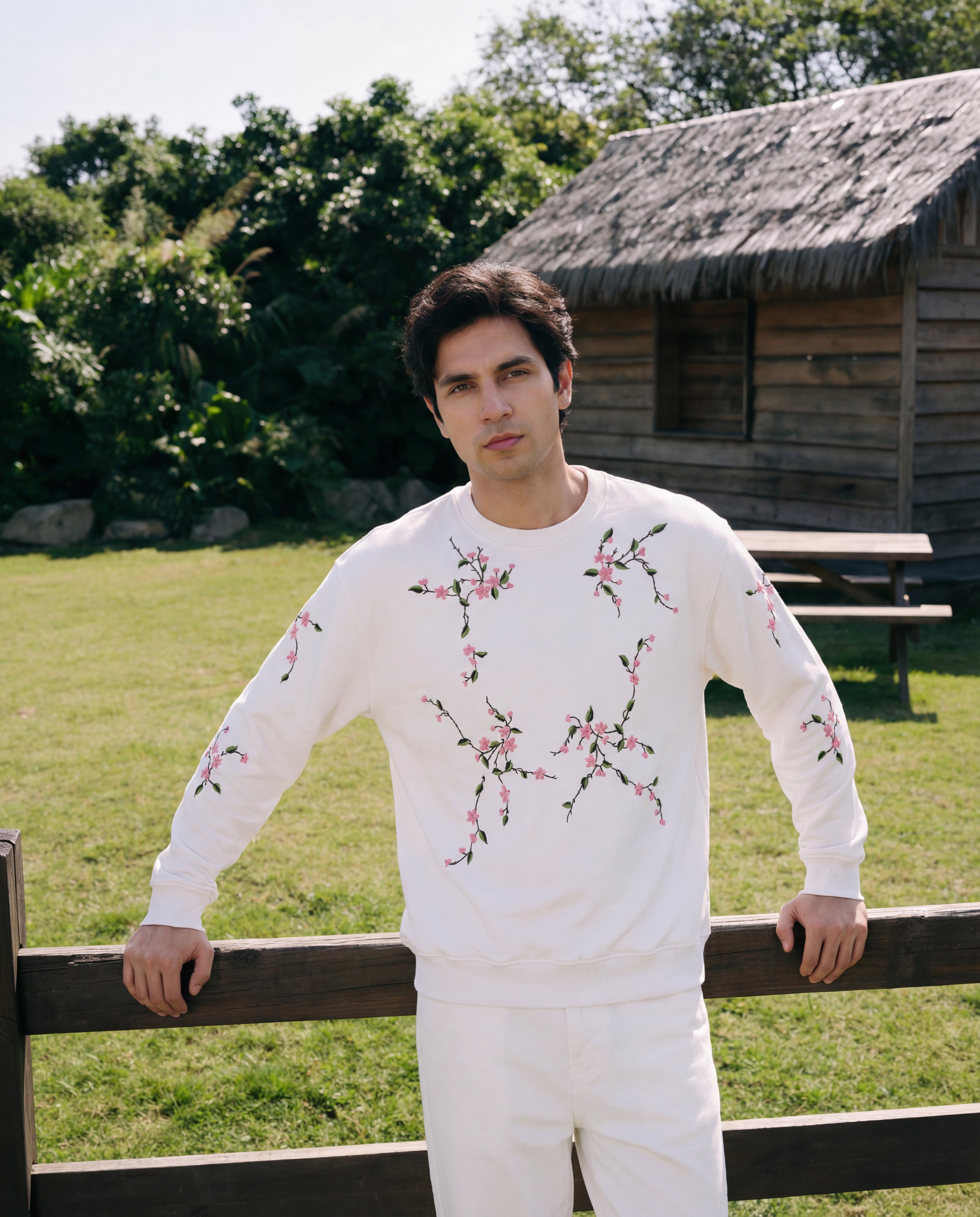 Man wearing a white outfit with floral patterns standing outdoors near a wooden cabin.