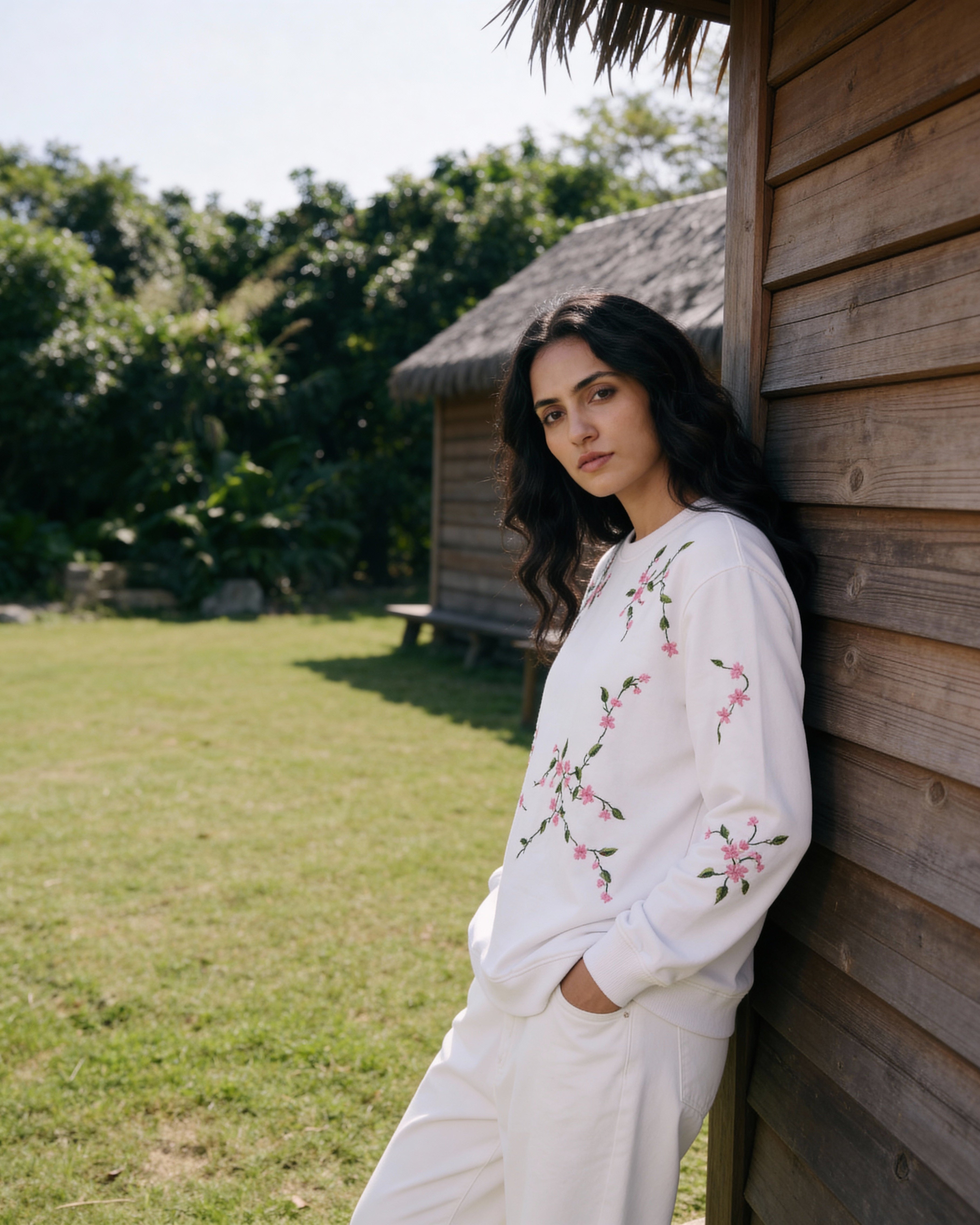 Woman in a white outfit with floral embroidery leaning against a wooden wall outdoors.