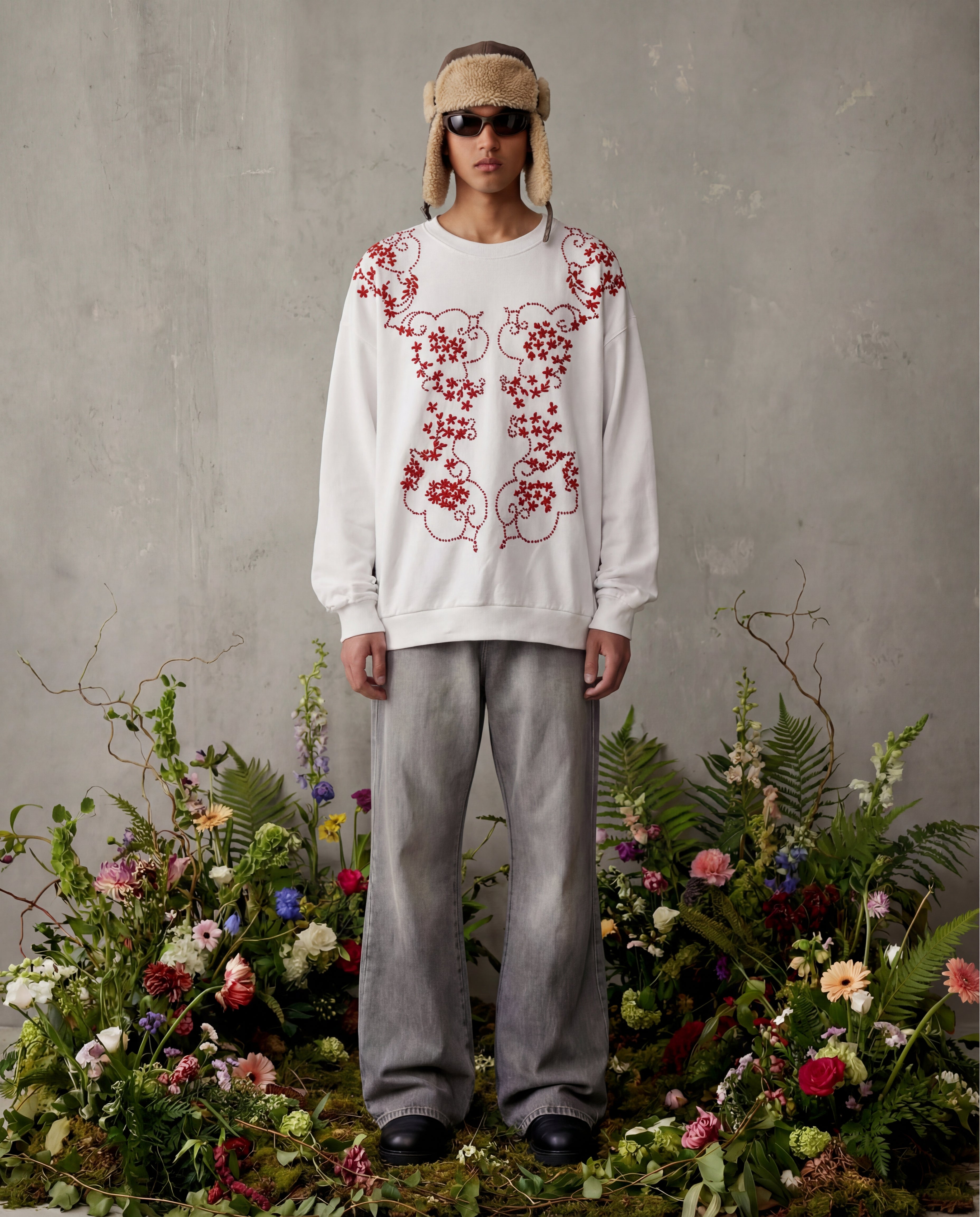 Person wearing a white sweatshirt with red floral embroidery and gray pants, standing among flowers and plants.