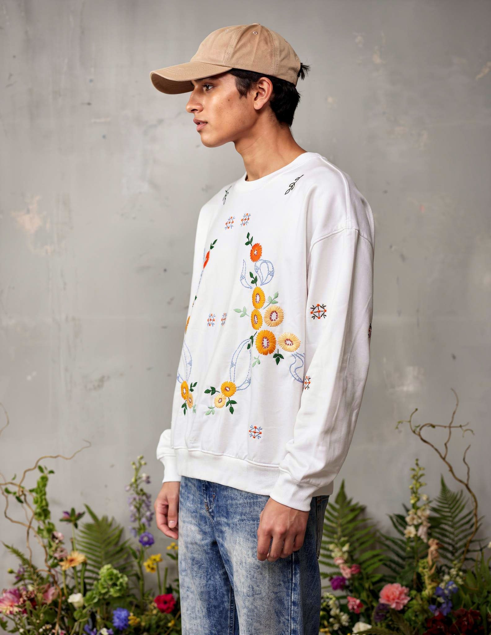 Person wearing a white sweatshirt with floral embroidery against a gray wall with plants.
