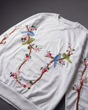 White sweatshirt with embroidered birds and floral branches on a dark background