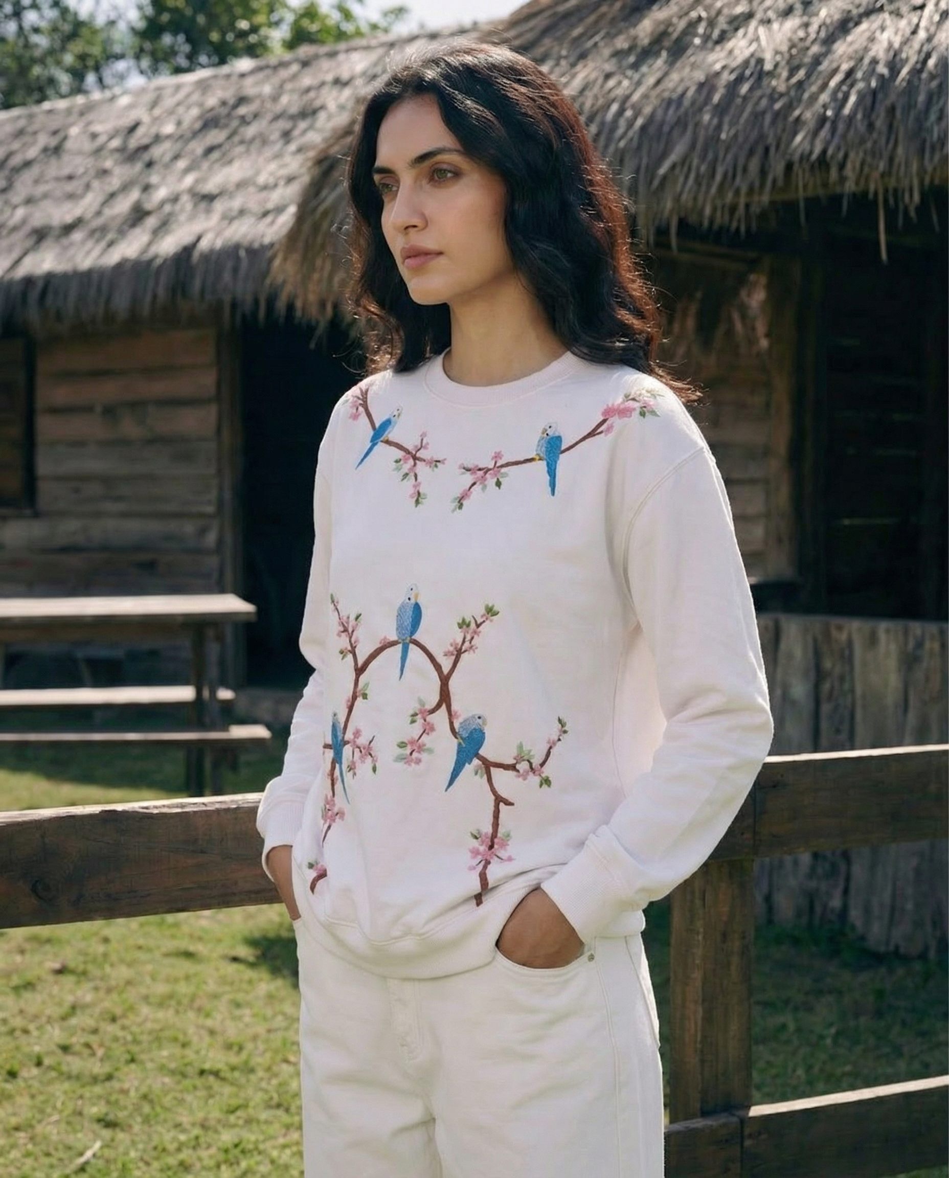 Woman wearing a white sweatshirt with bird hand embroidered designs outdoors near rustic wooden structures.