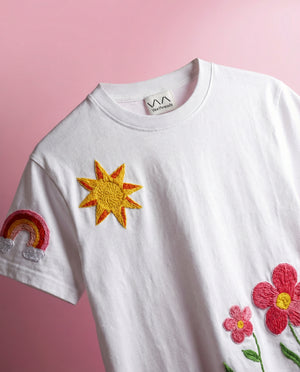White t-shirt with embroidered sun, rainbow, and flower designs on a pink background