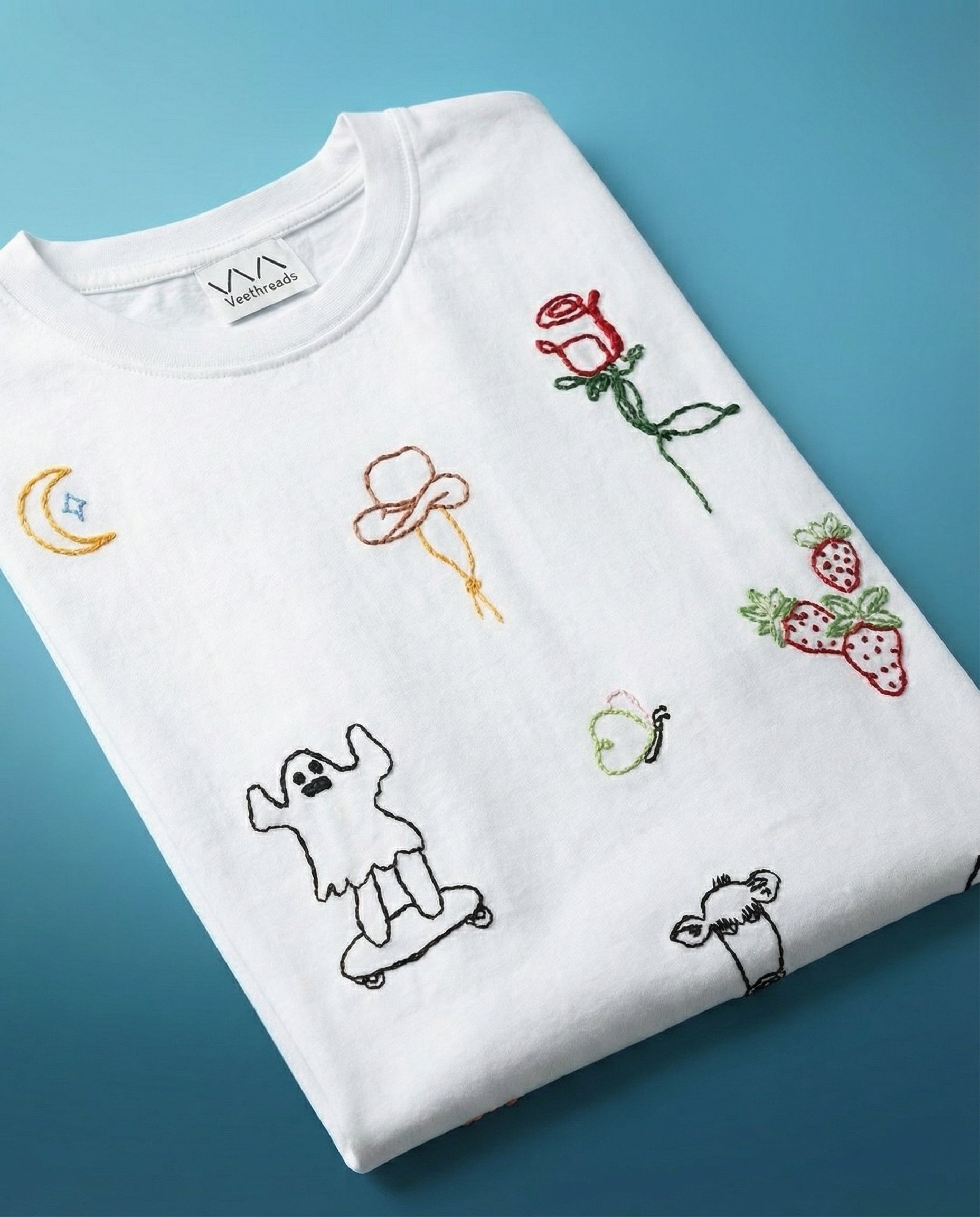 Random doodle embroidered tshirt — white cotton tee with hand-embroidered doodle designs including butterfly, moon, ghost and cow motifs on blue background