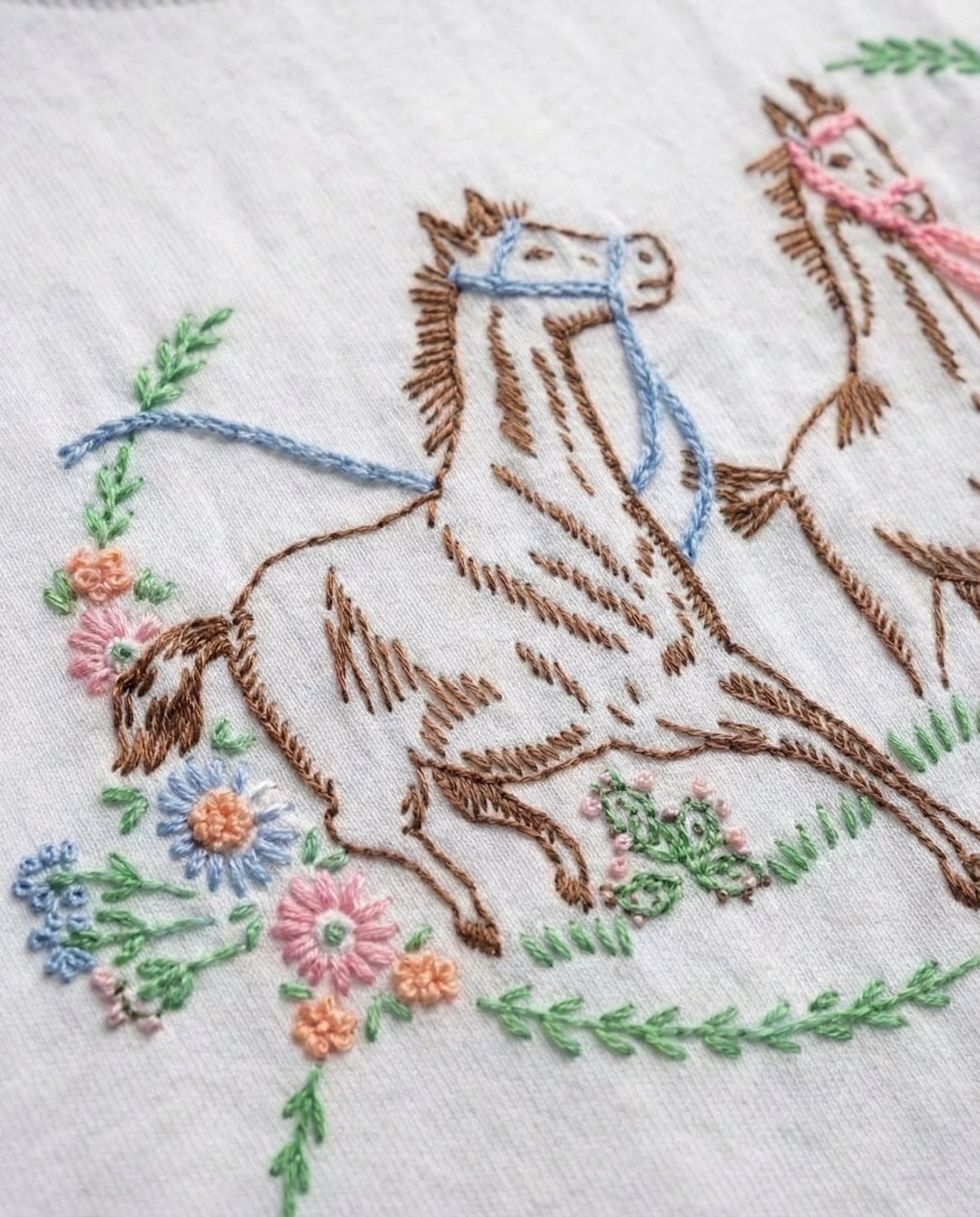 Animal embroidery designs horse — close-up of hand-embroidered horse with floral wreath elements on white cotton fabric