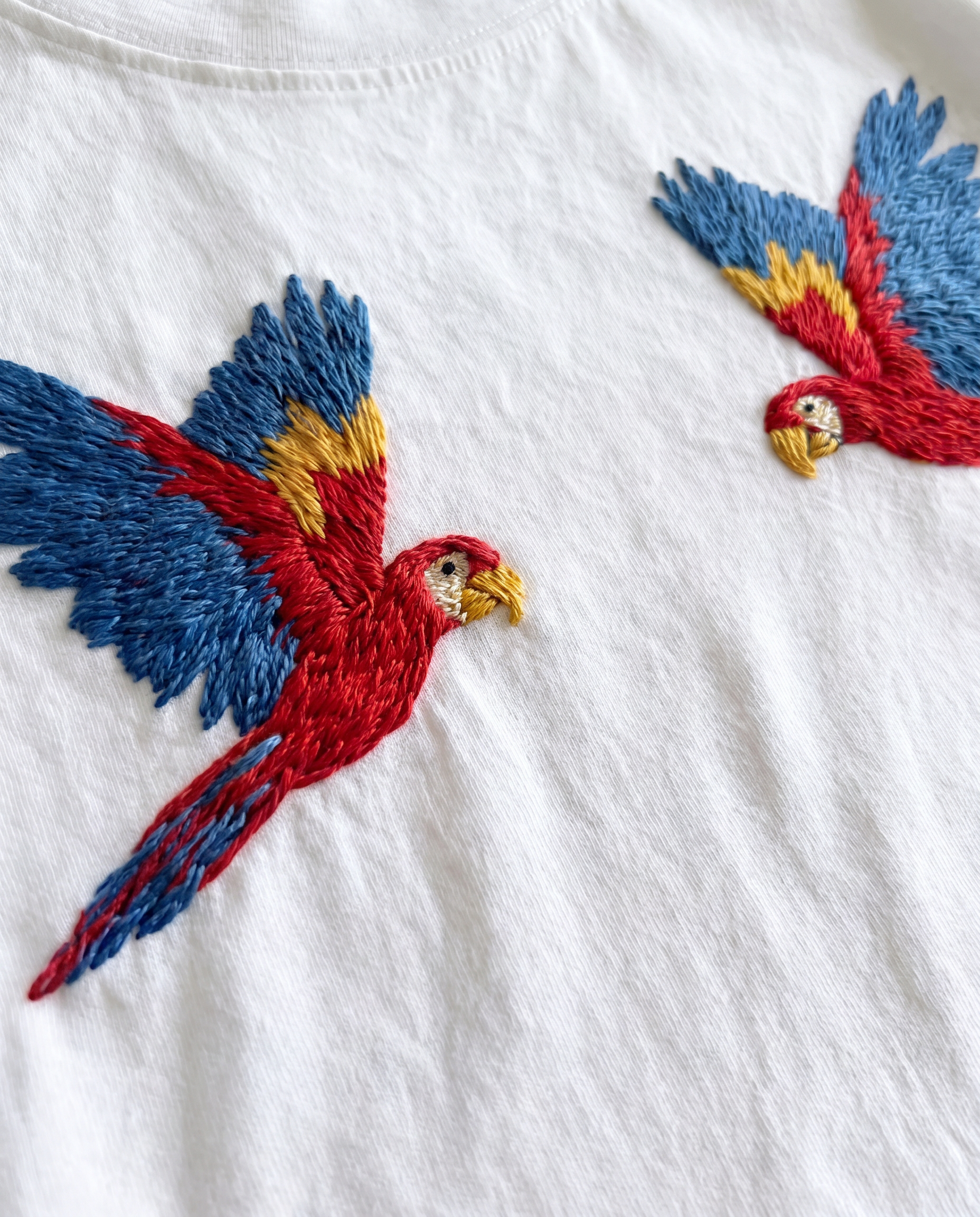 close-up of two parrots — scarlet macaws — hand-embroidered in red, blue and gold thread on a white cotton t-shirt, showing the feather thread work.