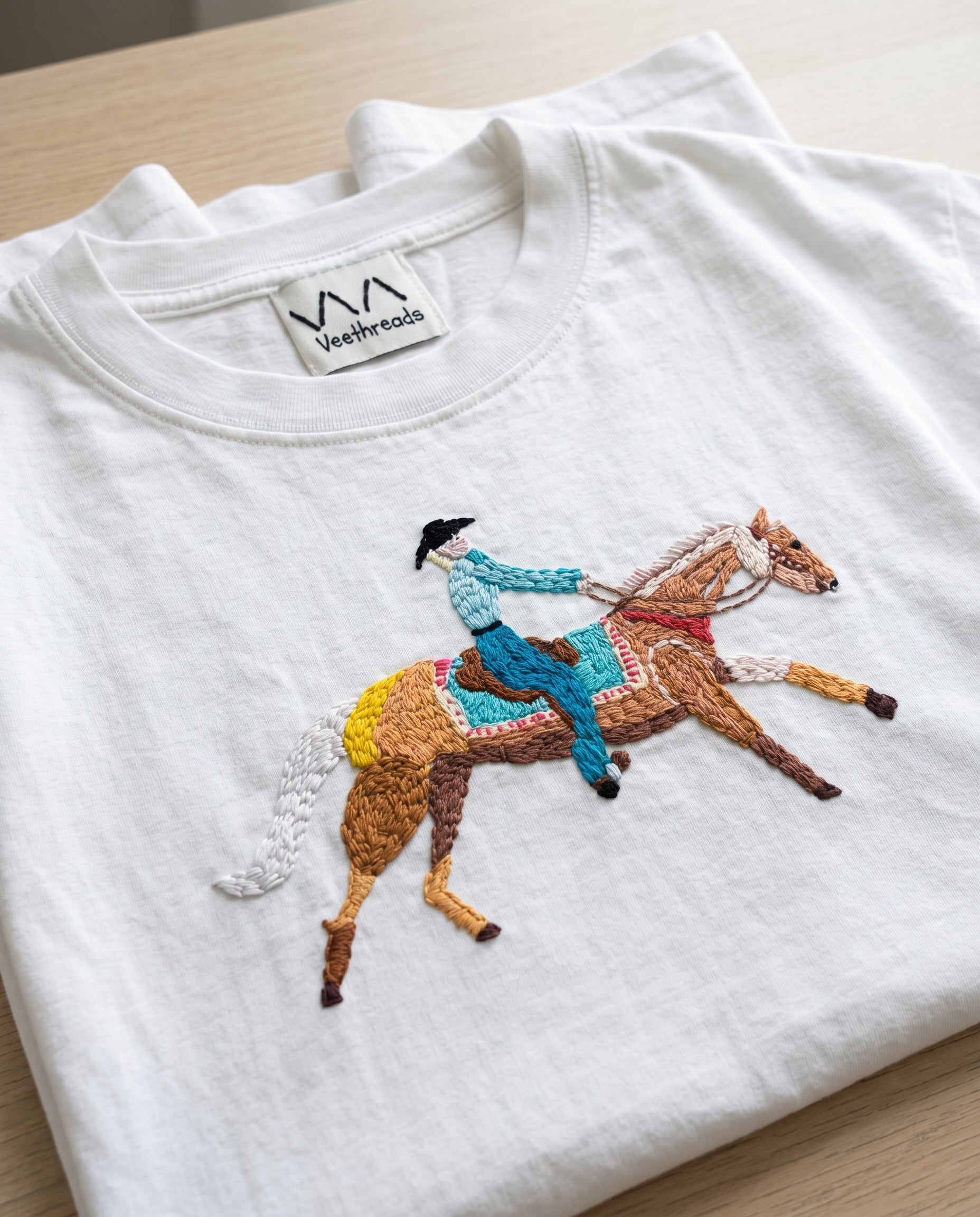 Close-up of a hand-embroidered cowboy and horse on a folded white cotton t-shirt — Vee Threads
