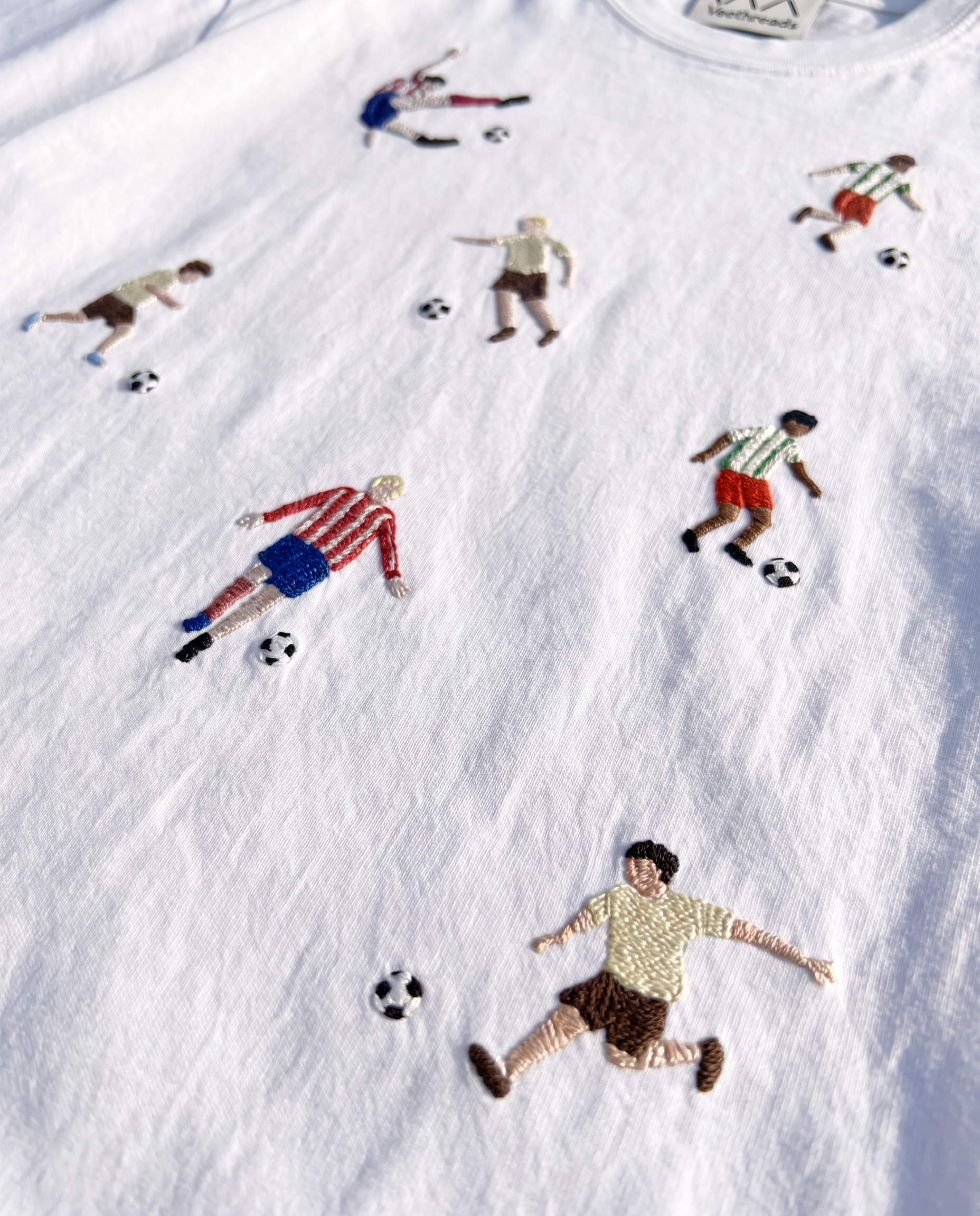 White cotton tee with footballers in striped and solid kits hand-embroidered across the front, each with a soccer ball, by Vee Threads