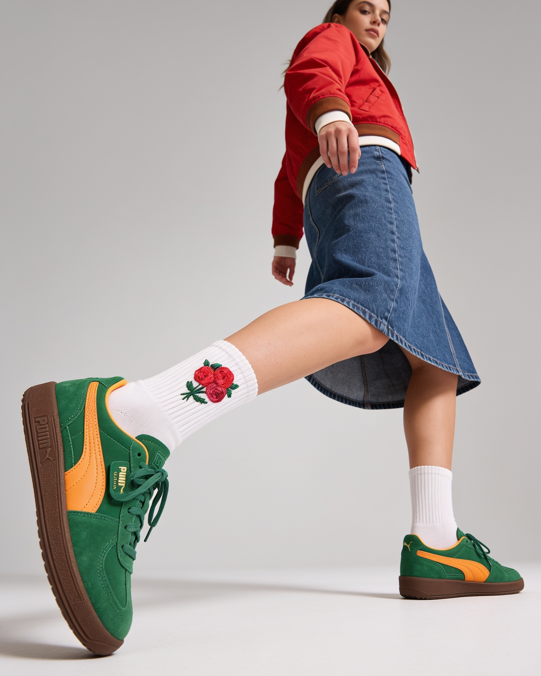 A model wearing green Puma sneakers with yellow side stripes, white crew socks featuring a red rose graphic, a blue denim skirt, and a red jacket, captured in a dynamic pose against a neutral gray studio background.