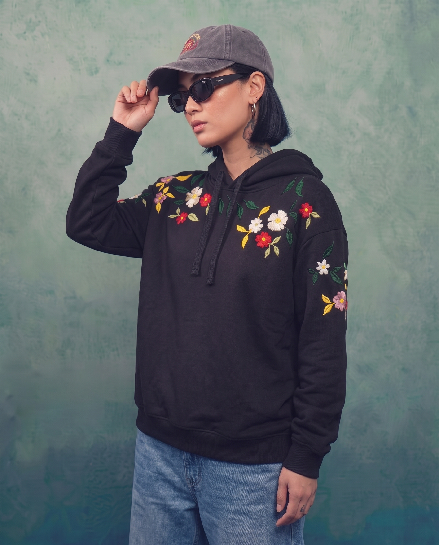 Woman wearing the black floral embroidered hoodie with a cap and sunglasses, arm raised