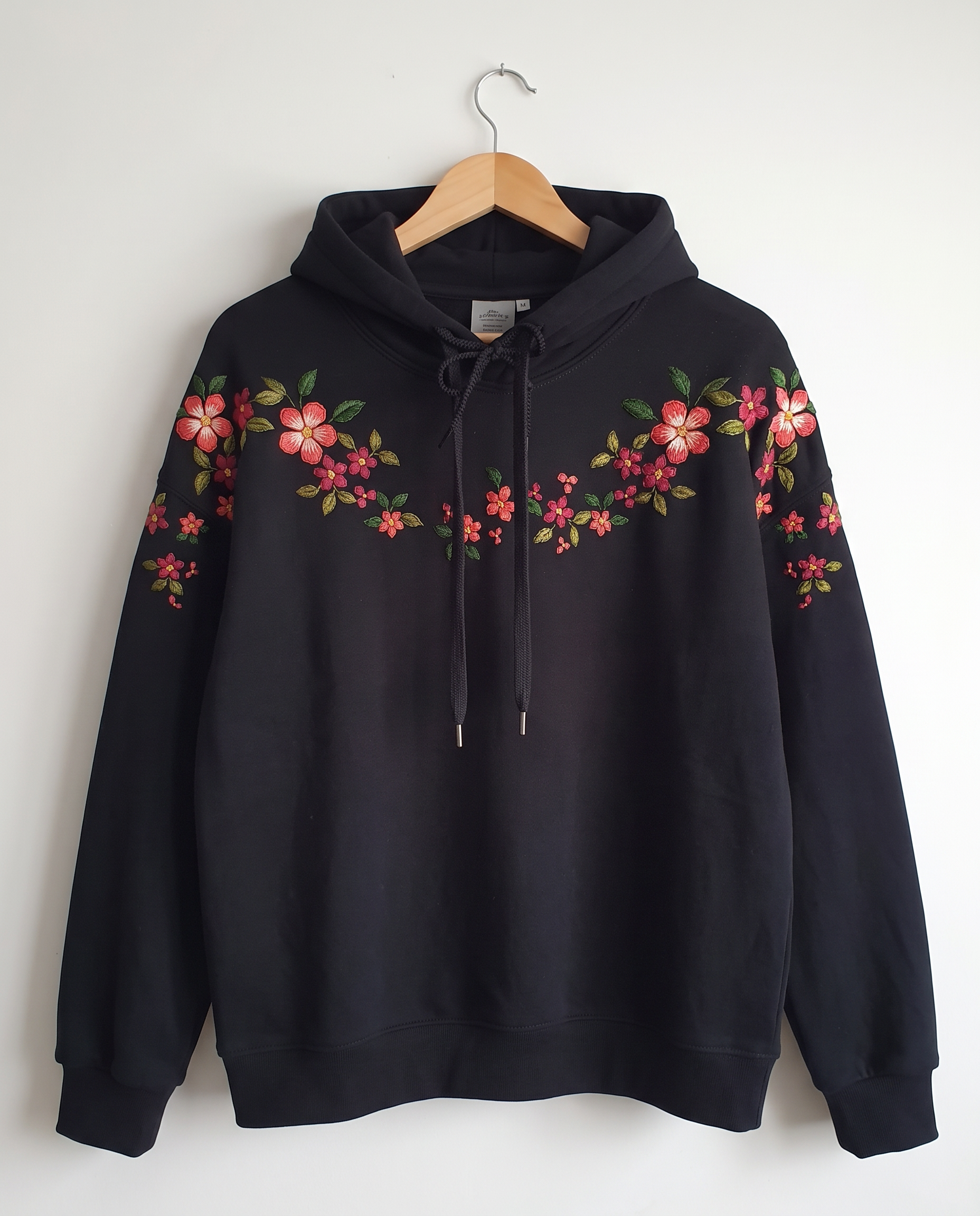 Black cotton fleece hoodie on a hanger with coral and pink blossoms hand-embroidered across the shoulders
