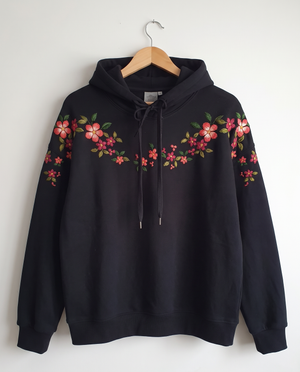 Black cotton fleece hoodie on a hanger with coral and pink blossoms hand-embroidered across the shoulders