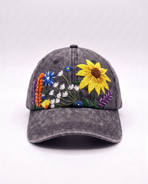 Charcoal acid-wash denim cap, front view, with a hand-embroidered wildflower garden around one large yellow sunflower
