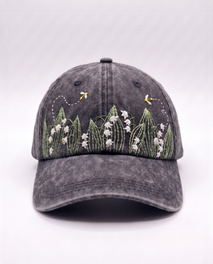 Charcoal acid-wash denim cap, front view, with white lily of the valley bells and two embroidered bees above them