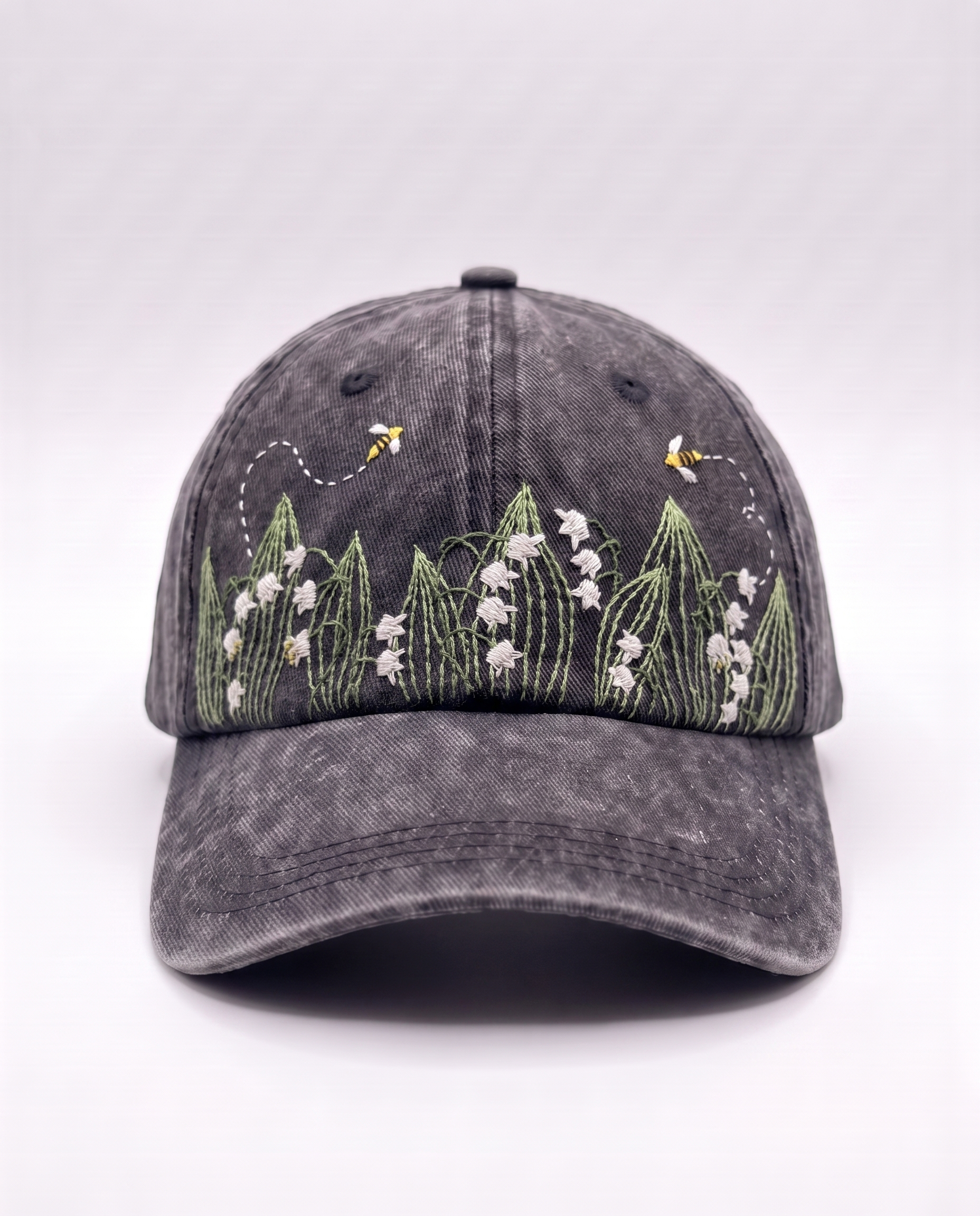 Charcoal acid-wash denim cap, front view, with white lily of the valley bells and two embroidered bees above them