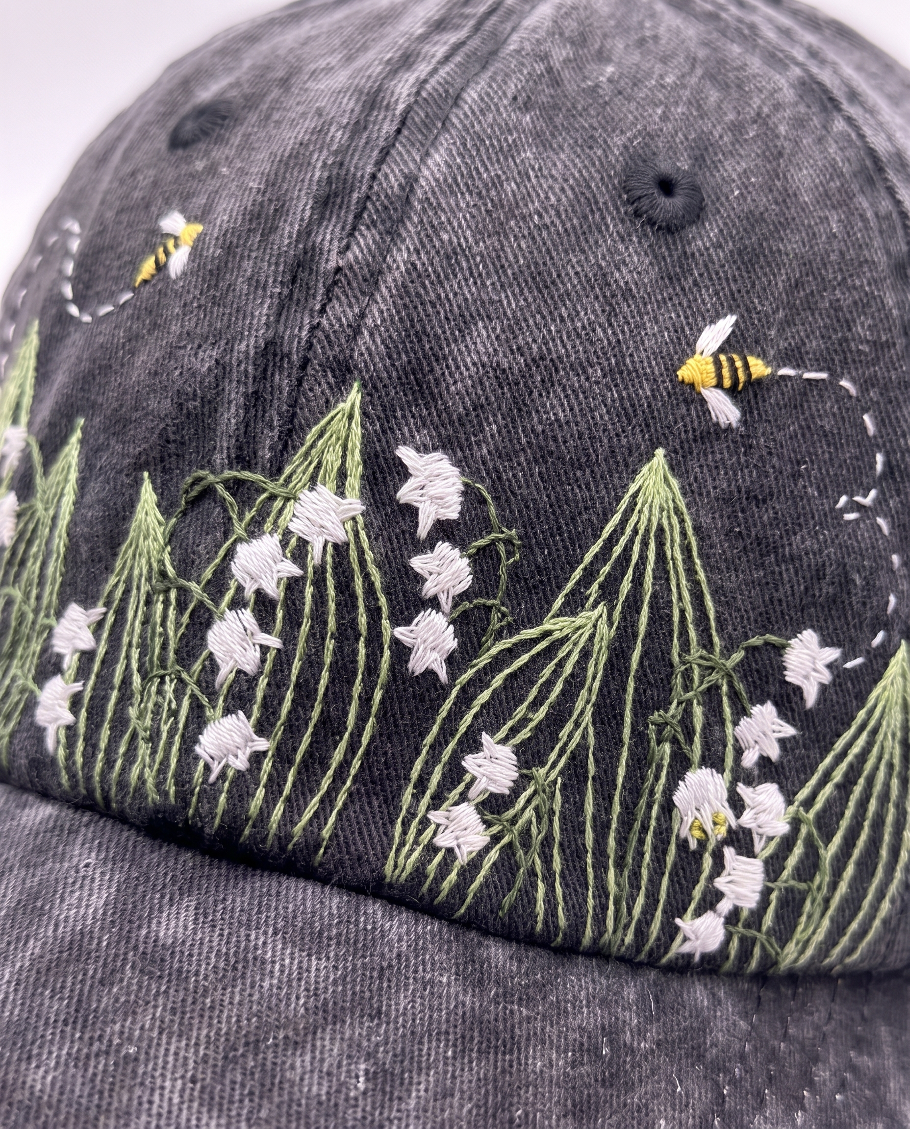 Detail crop of the embroidered band, white bell flowers on green blades with a bee and its dashed trail above