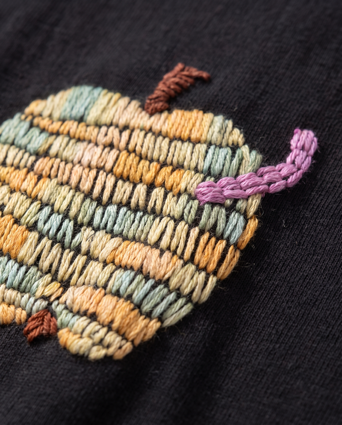 Close-up of the embroidered apple in rows of sage, apricot and cream stitches with a lilac chain-stitch worm