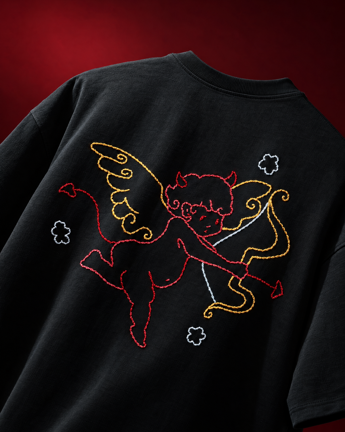 A close-up of a black t-shirt featuring a large embroidered graphic of Cupid on the back. The embroidery depicts Cupid in red and yellow, holding a bow, with small white cloud motifs scattered around.