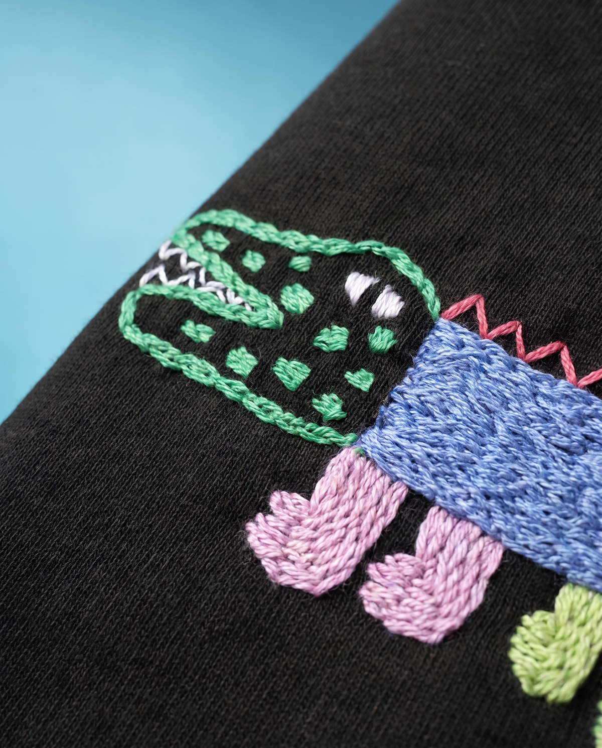 Close-up of the crocodile embroidery, green chain-stitch head with white teeth, blue textured body and pink stitched legs