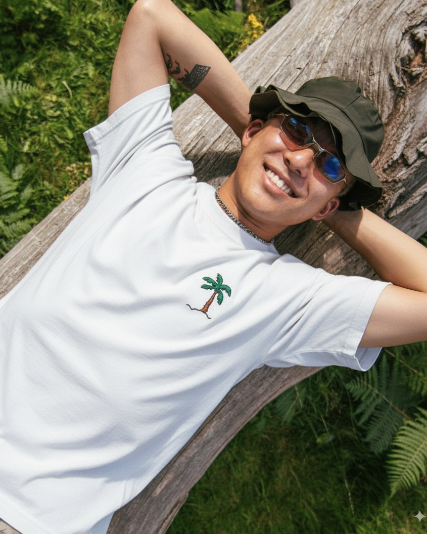 Person wearing a white shirt with a palm tree design embroidered , sunglasses, and a cap, leaning against a wooden log outdoors.